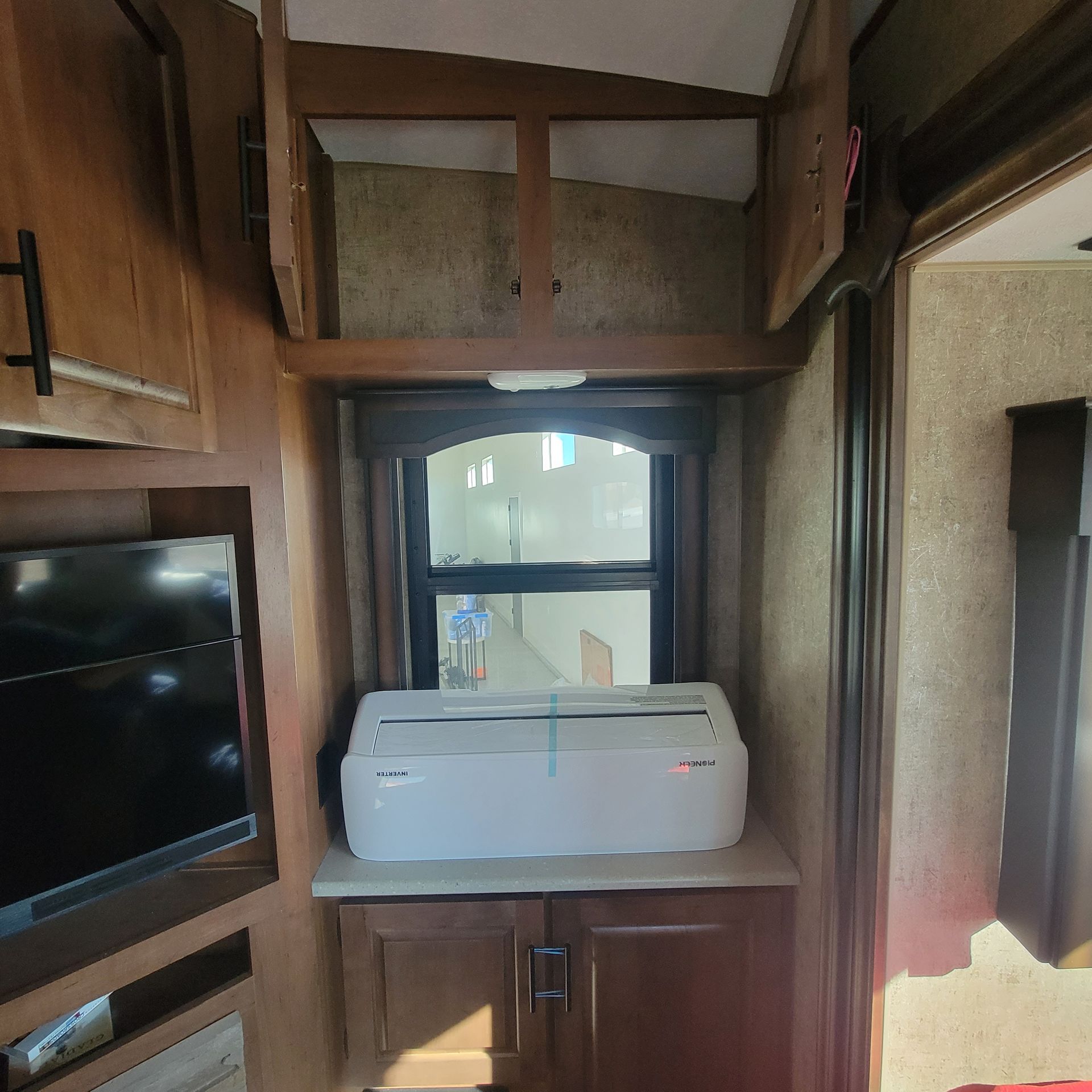 RV interior: window with a printer on a cabinet, surrounded by brown wood cabinets and a TV.