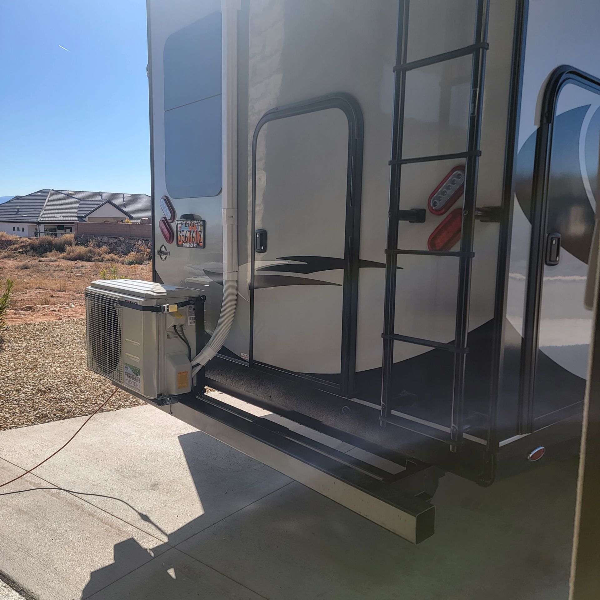 Rear of a white RV with a mounted air conditioner, ladder, and access door. Set on concrete in a sunny location.