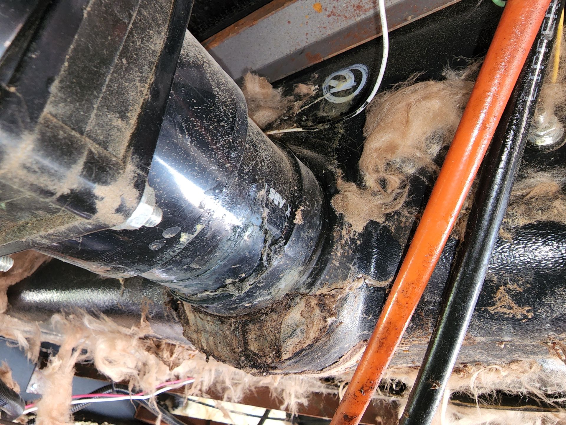 Black plumbing pipes with dirt and insulation in a cramped space. An orange pipe runs alongside.