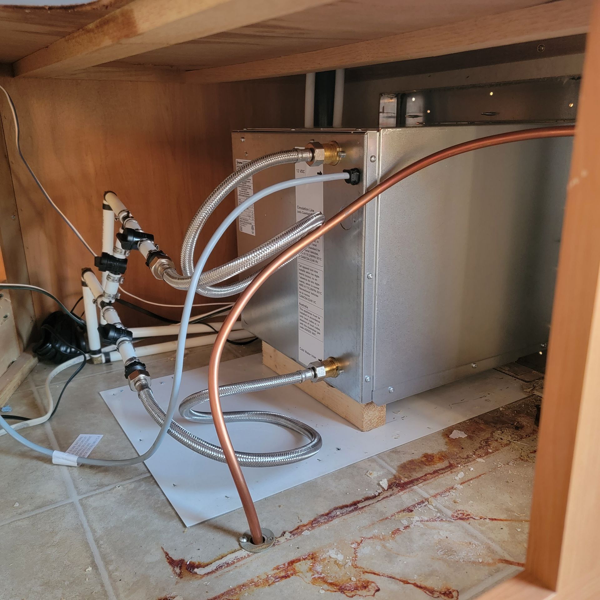 Underside of cabinet with plumbing, a metal water heater, and connecting pipes.