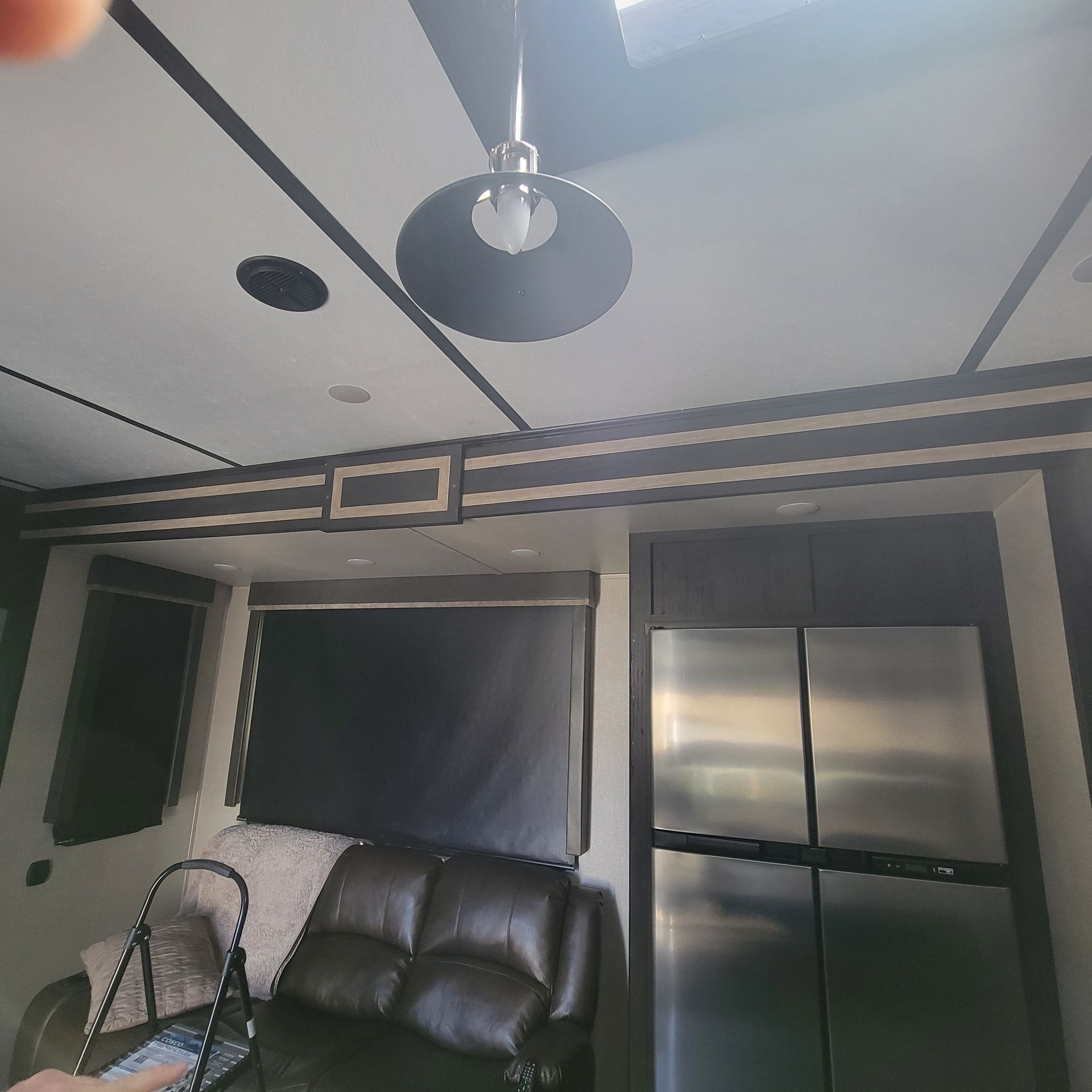Interior view of an RV living area: black leather couch, stainless steel fridge, overhead lighting, black window shades.