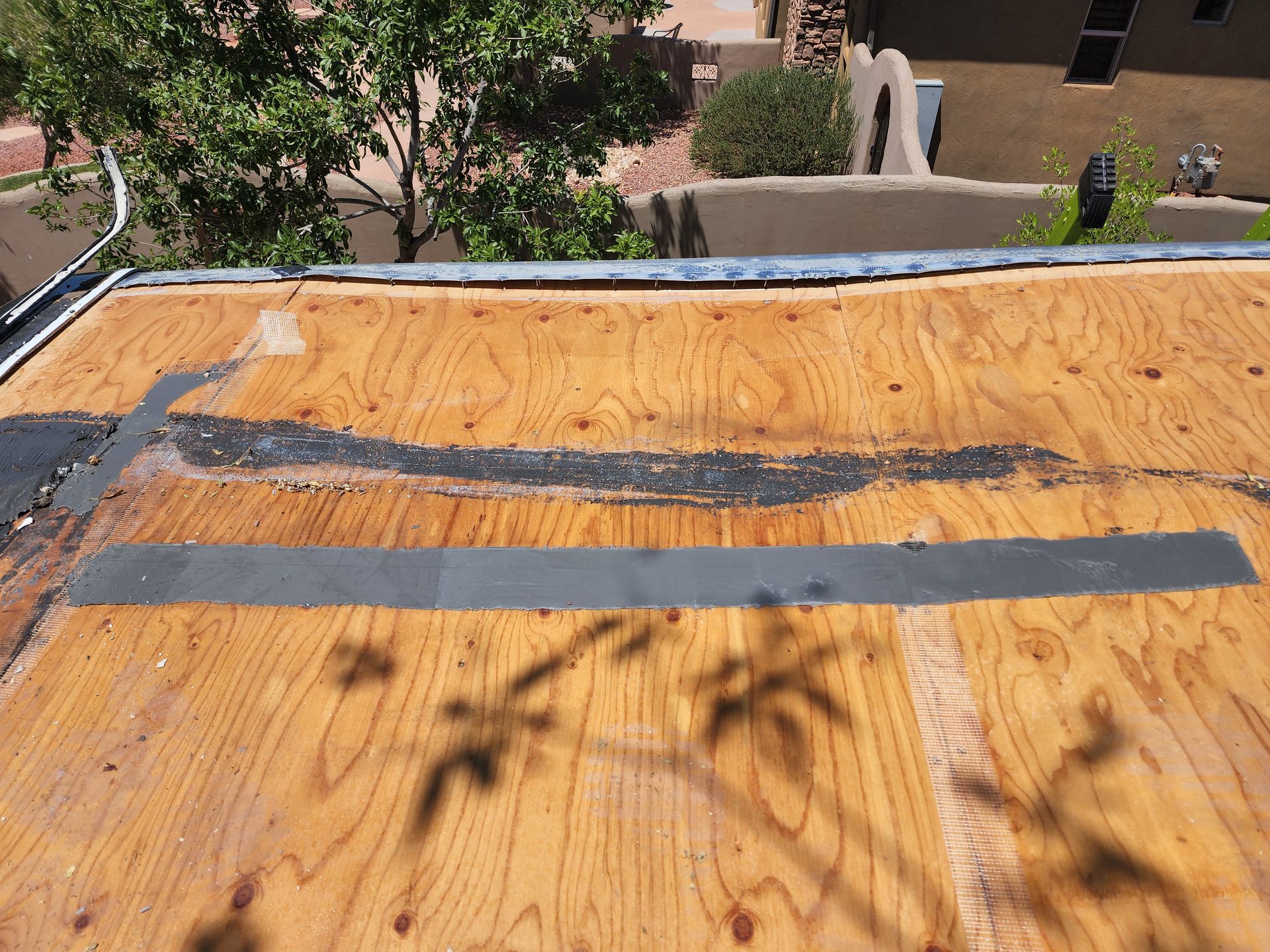 Wooden roof with patches, featuring a gray strip and black sealant, with building and trees in the background.