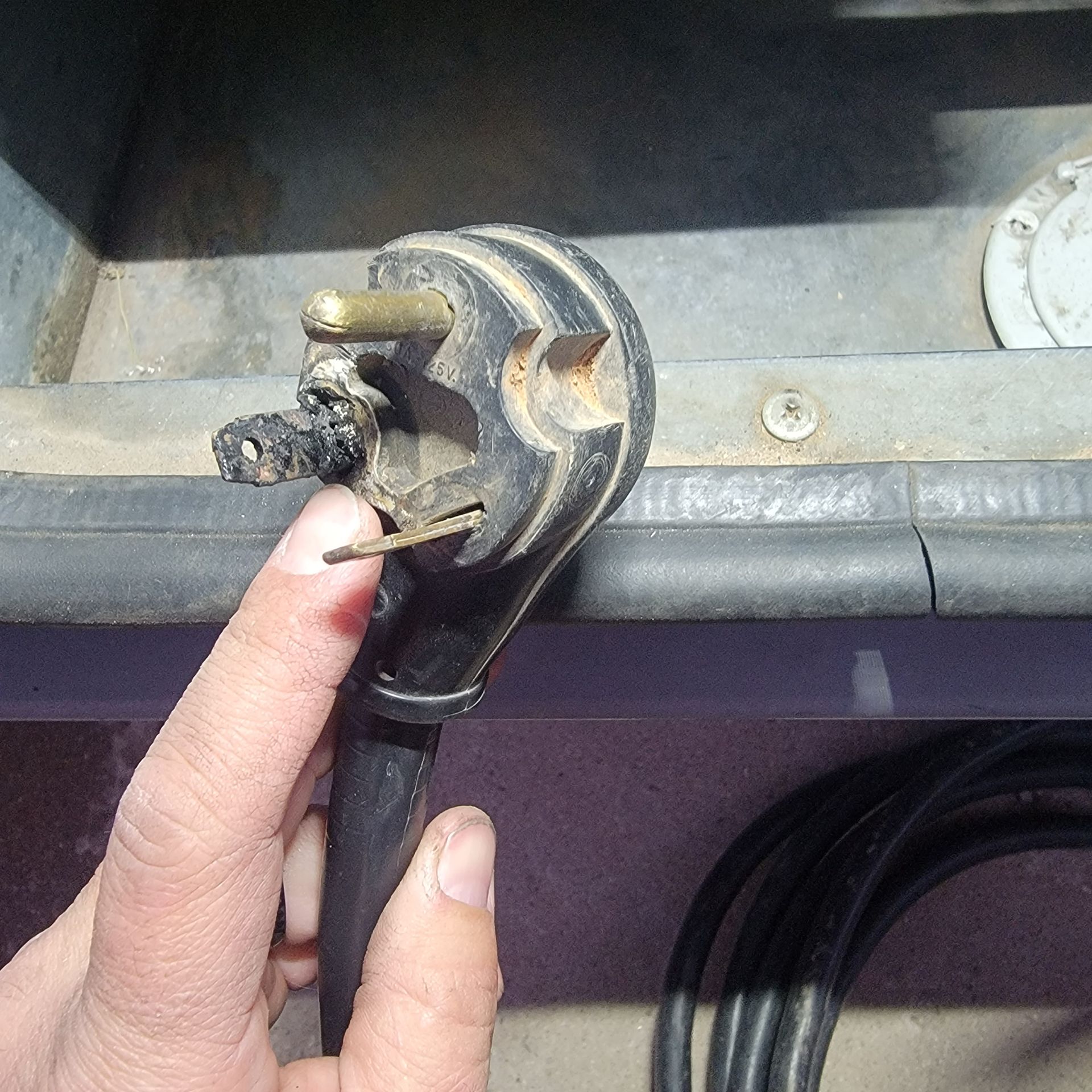 Hand holding a damaged black electrical plug with three prongs, near a metal surface.