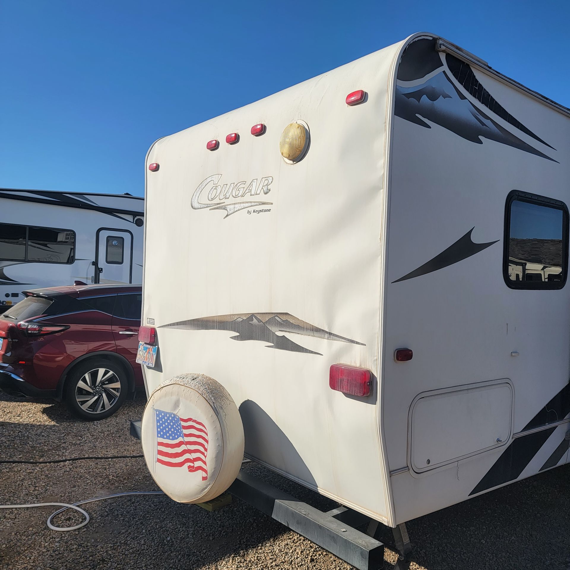 White Cougar travel trailer with spare tire cover bearing the American flag. Red car and another RV in background.