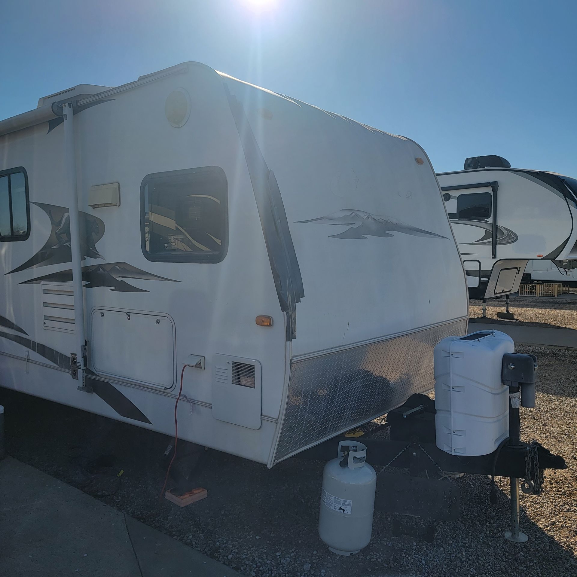 White RV trailer with propane tank in a gravel lot on a sunny day.