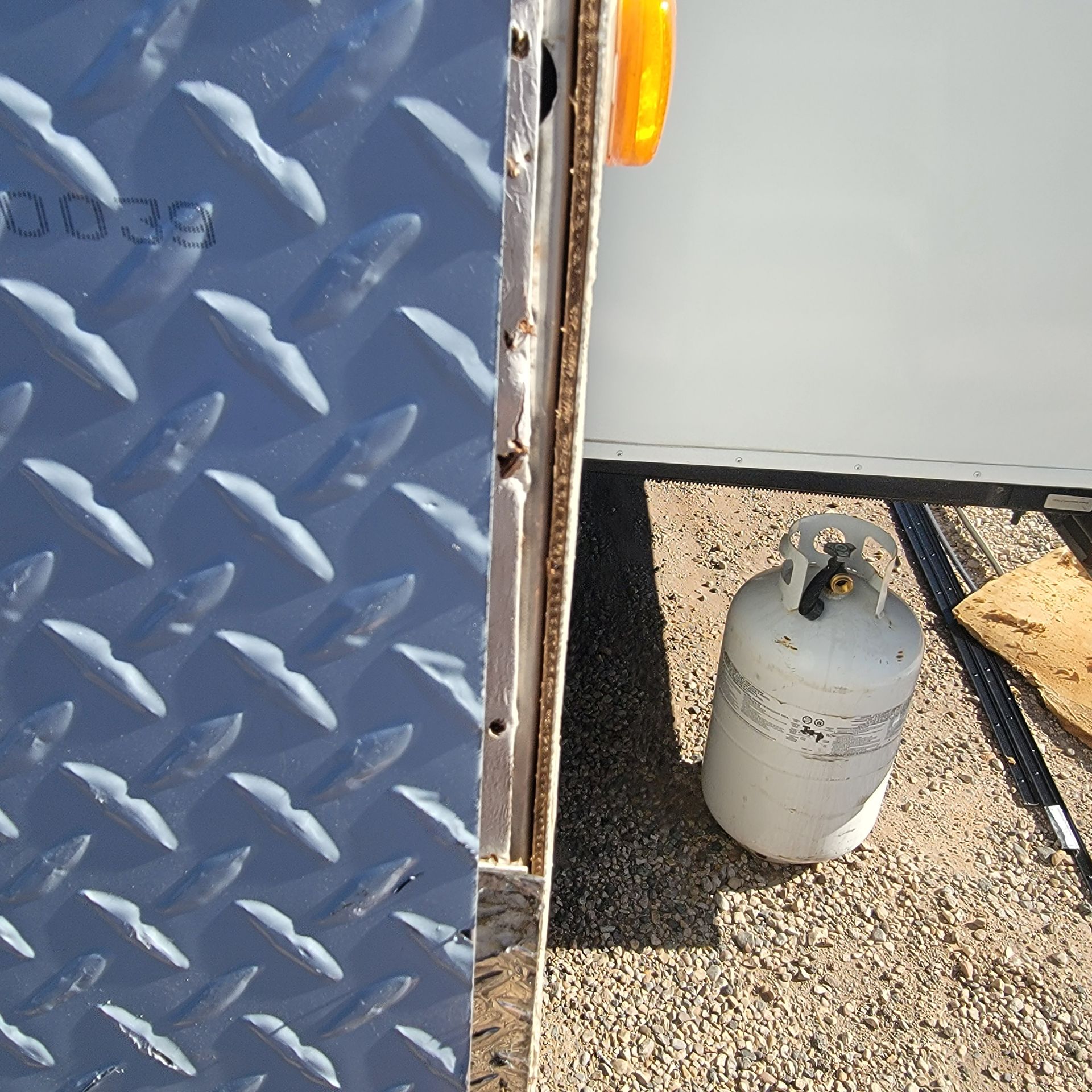 Blue diamond plate, metal trailer edge, propane tank, gravel ground, and amber light.