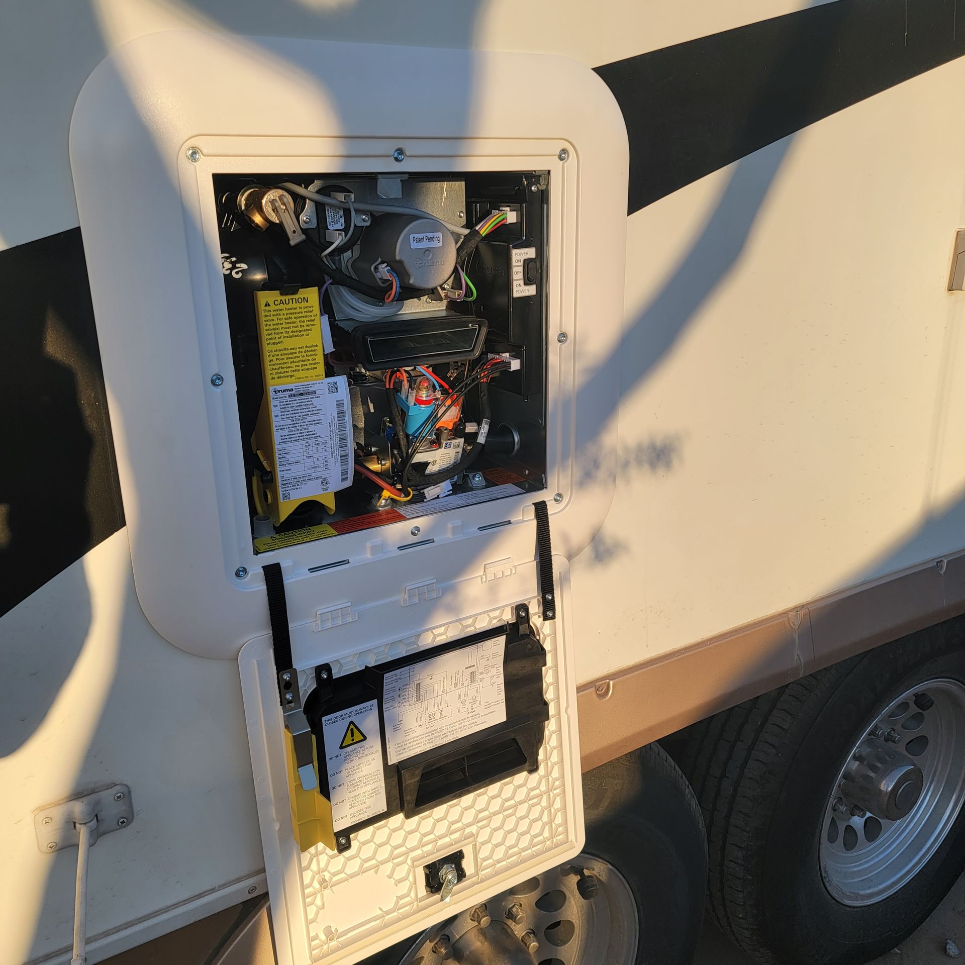 RV access panel open, revealing internal components and wiring. White exterior, black and brown accents, daytime.