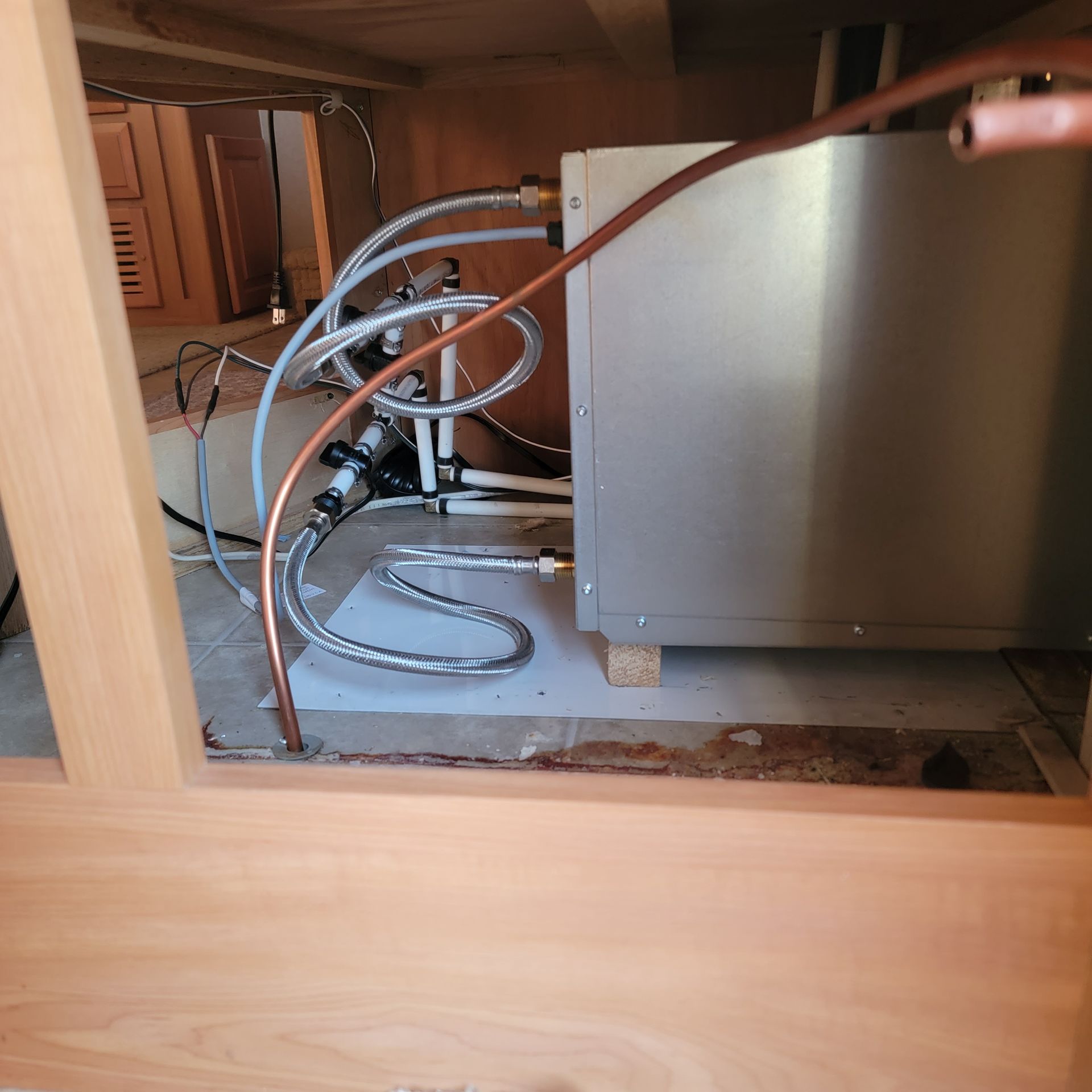 Plumbing and machinery under wooden cabinetry. Copper and silver tubes and a rectangular metal box.