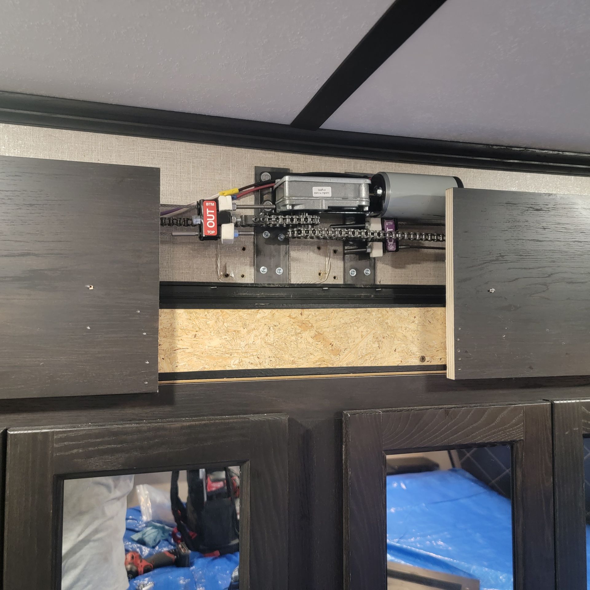 Motorized mechanism for sliding cabinet doors in a vehicle. Black cabinets, metal rails, and electric components are visible.