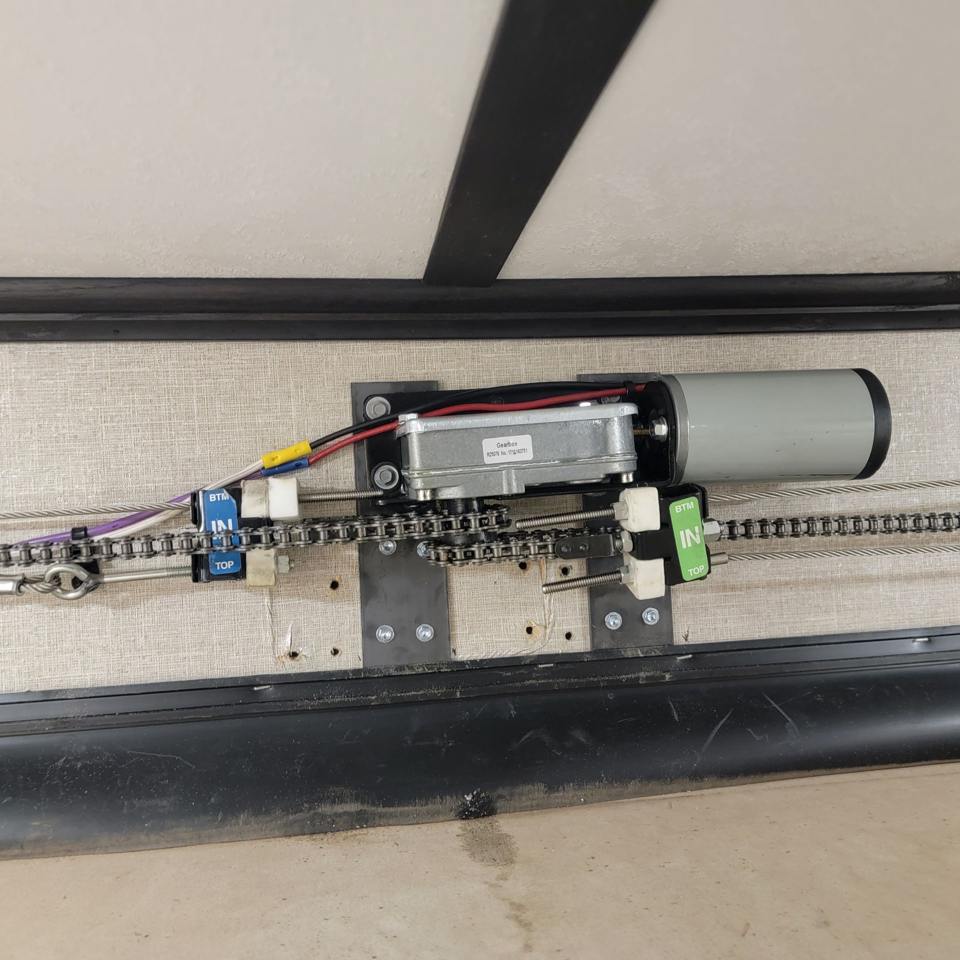 Motorized chain drive system mounted on a support structure; silver motor, black metal, chain, and wires.