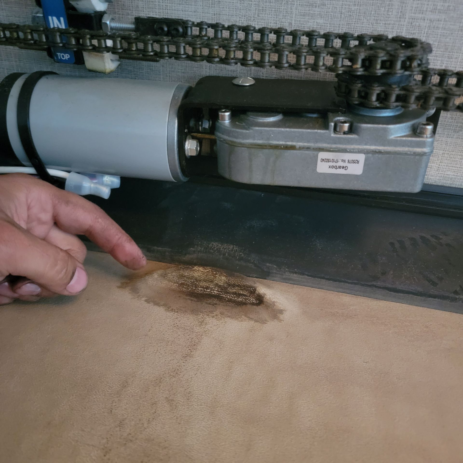 Person pointing at a brown, burned spot on a tan surface near a motor and chain.