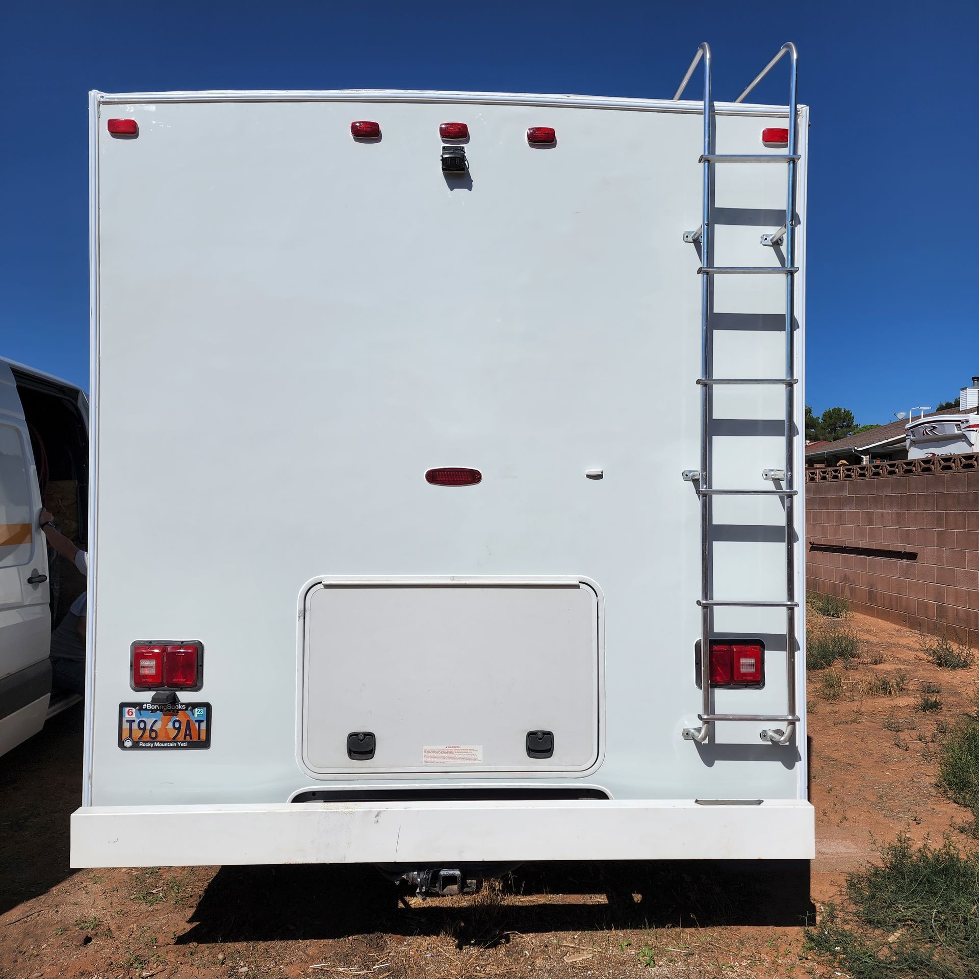 Rear view of a white RV with a ladder, taillights, and a storage door, parked outside on a sunny day.