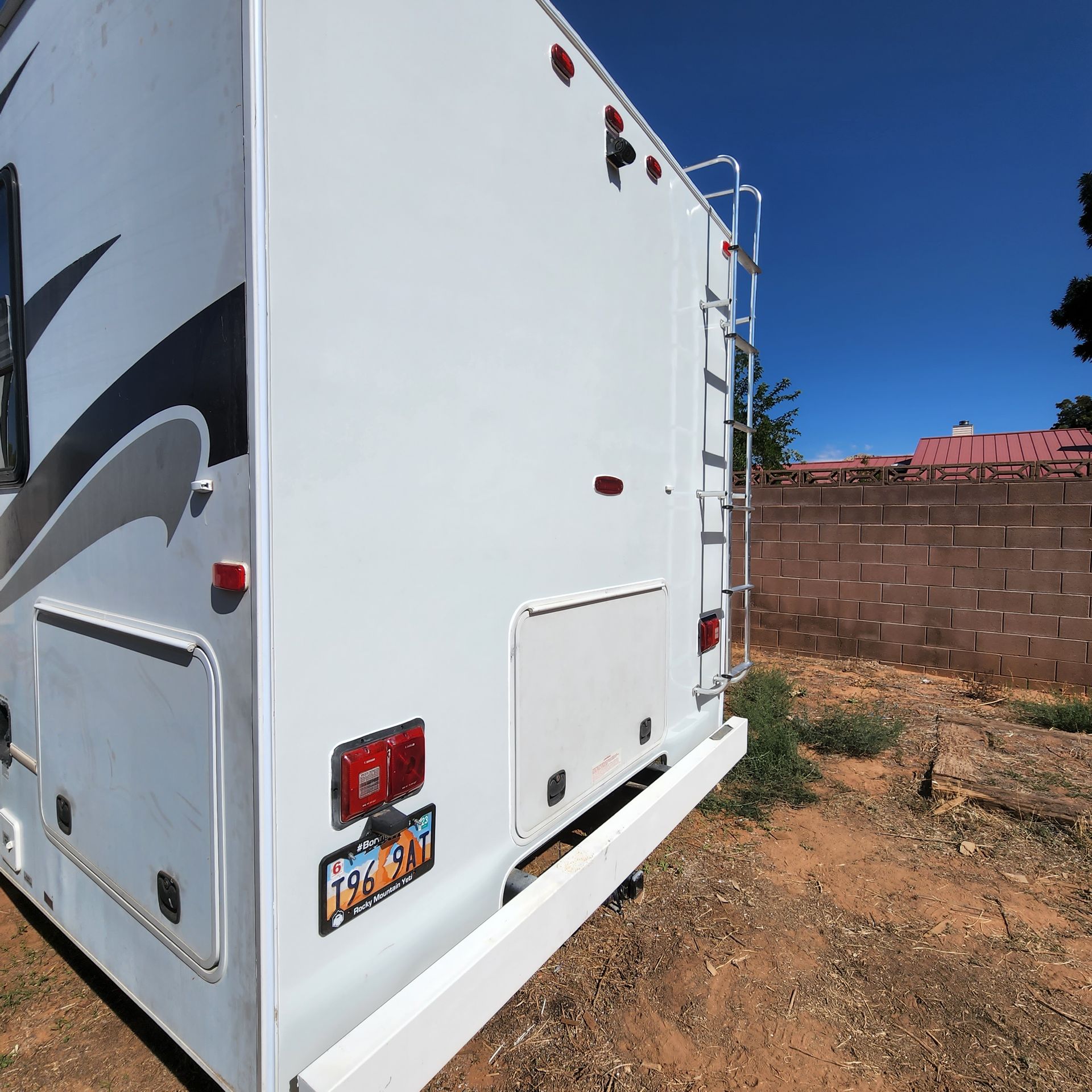 Rear side of a white RV parked outdoors; ladder, lights, and license plate visible.
