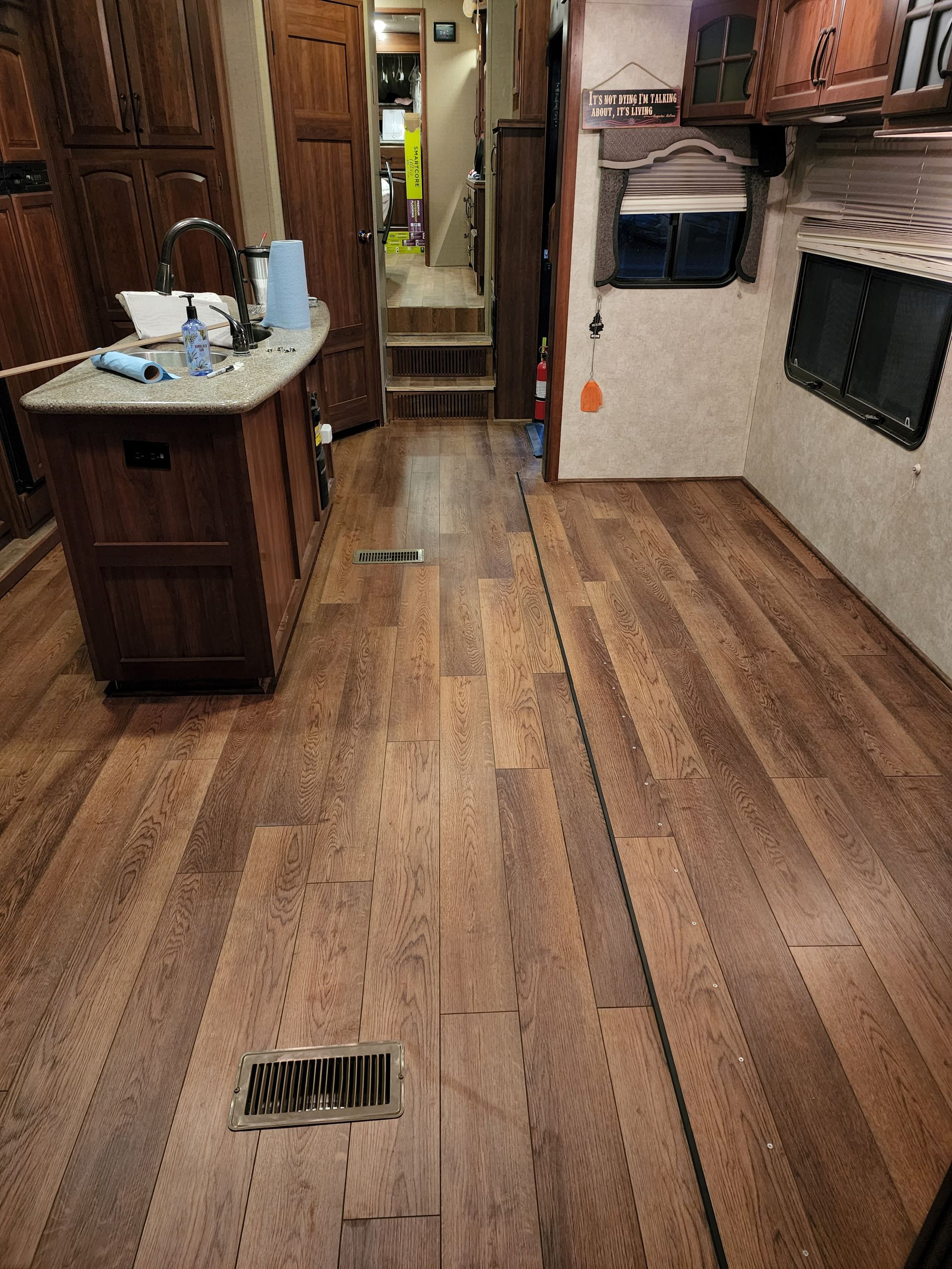 Interior view of a recreational vehicle with wood-look flooring, a kitchen island, and a hallway leading to the back.