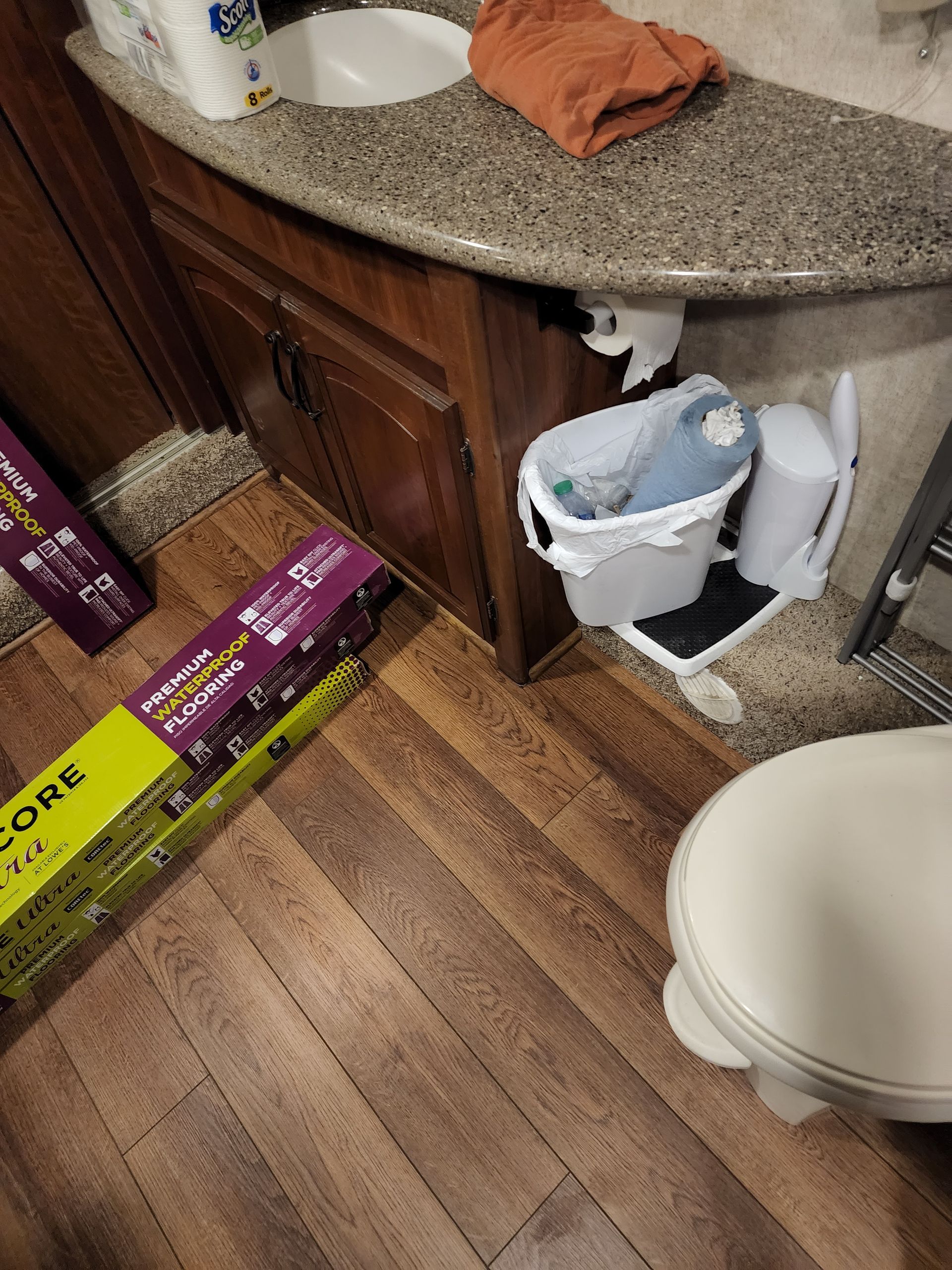 Bathroom with wood-look floor, countertop, vanity, toilet, trash can, and boxes of flooring.