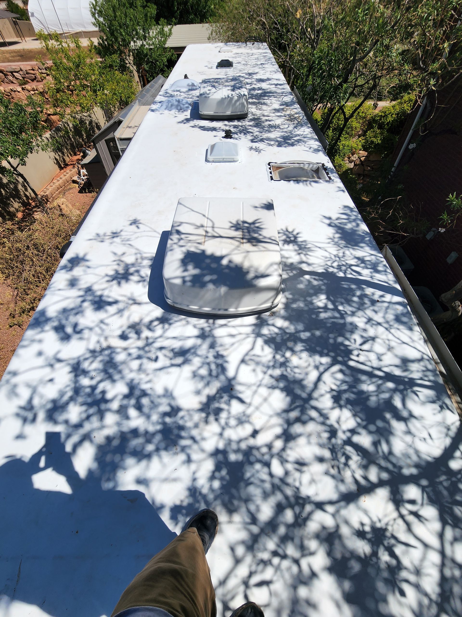 Overhead view of an RV roof with vents and a person's legs. Shadows of leaves cover the surface.