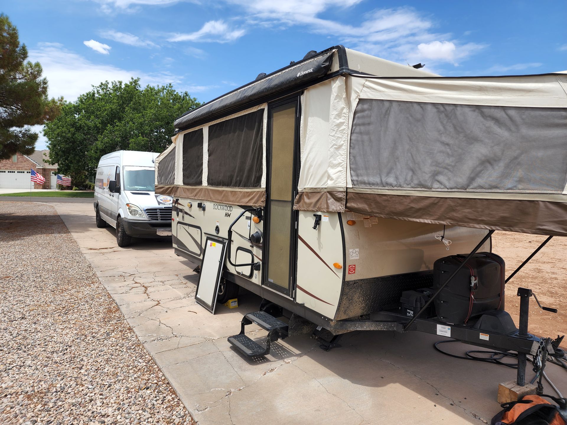 White van towing a tan and brown pop-up camper on a concrete driveway under a blue sky.