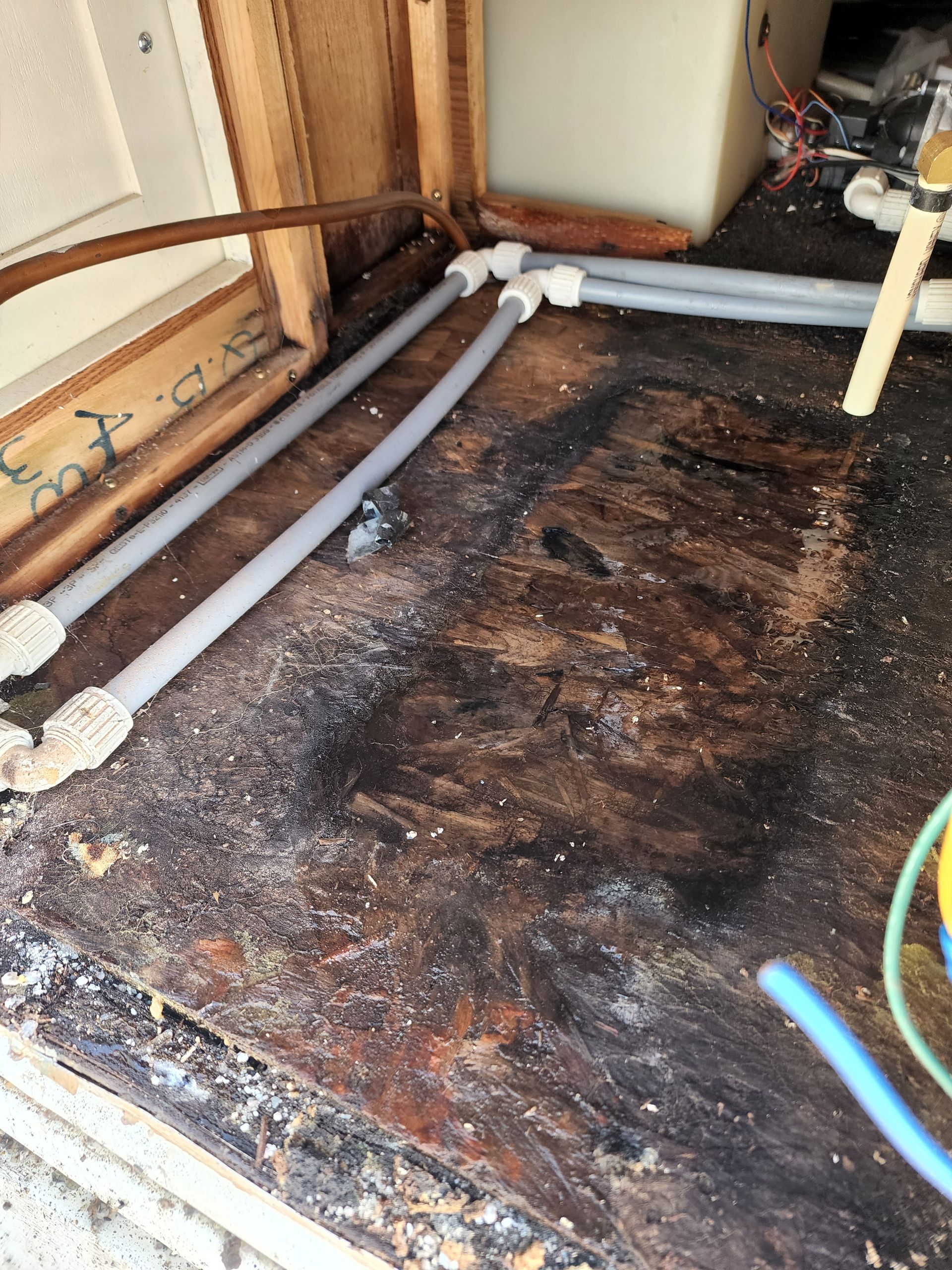 Damaged wooden floor with gray pipes and white connectors near a wooden frame.