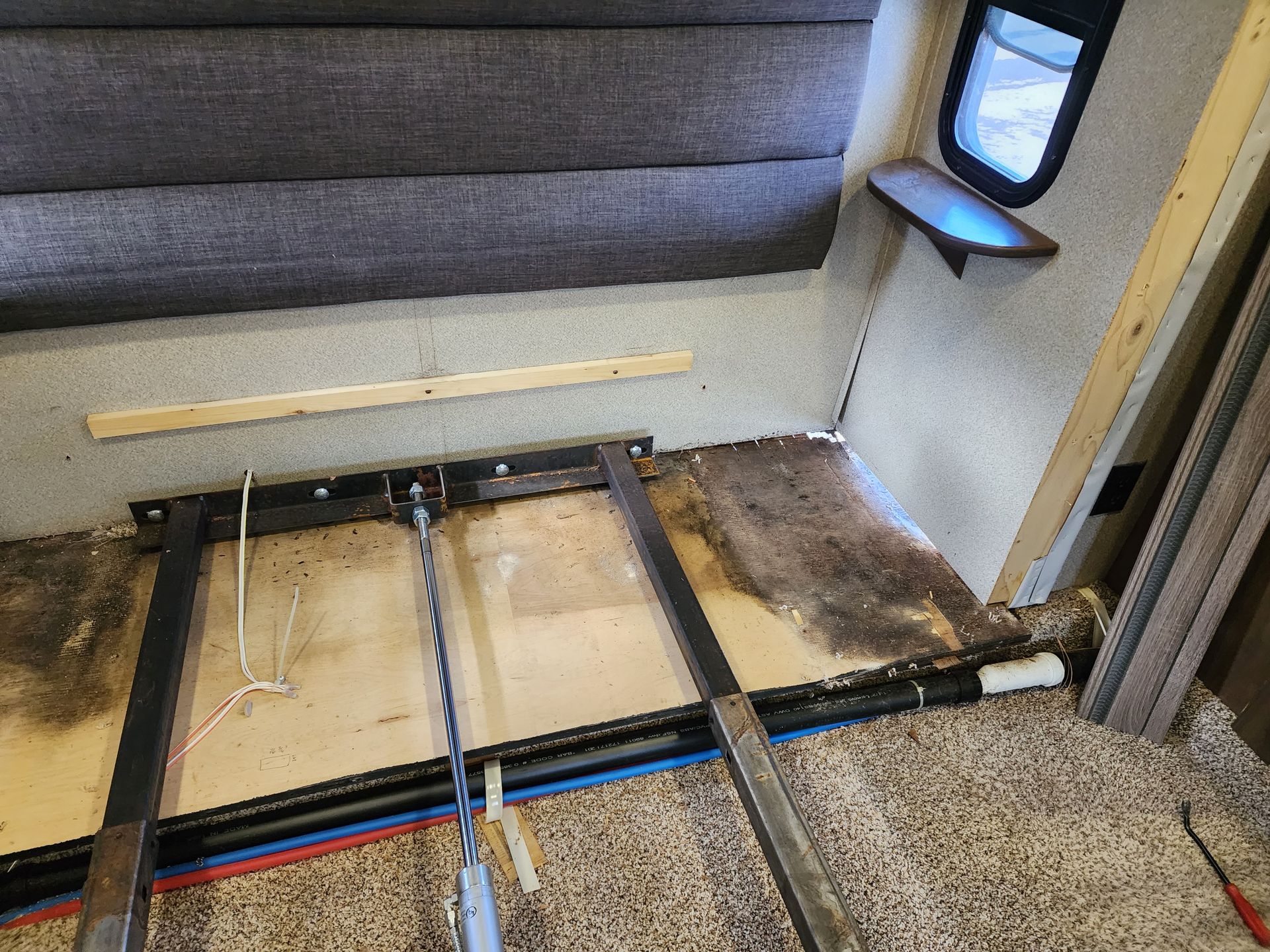 Interior of an RV bedroom under renovation, showing exposed floor, metal frame, and water pipes.