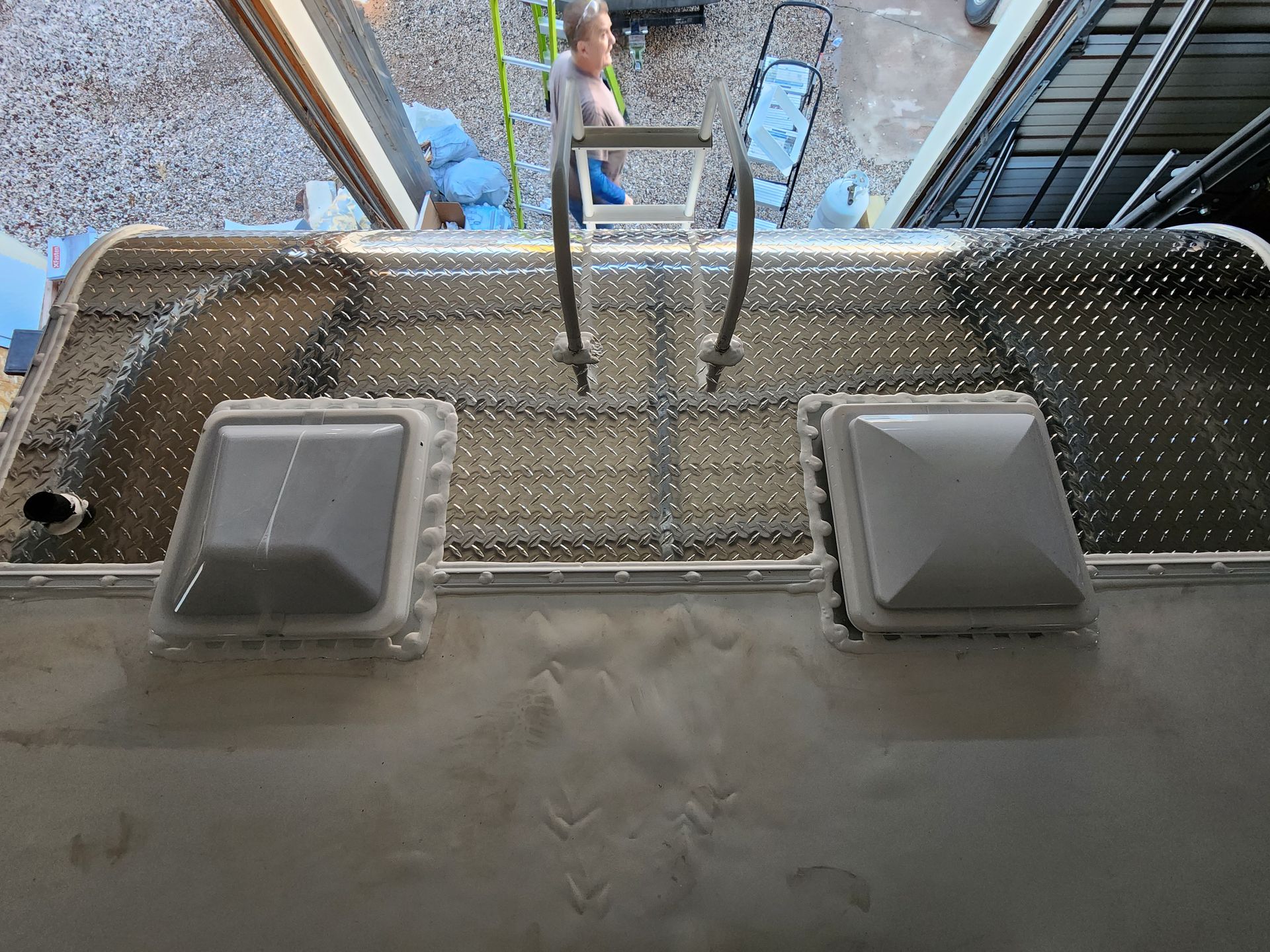 Silver RV roof with diamond plate, vents, ladder, person in background.