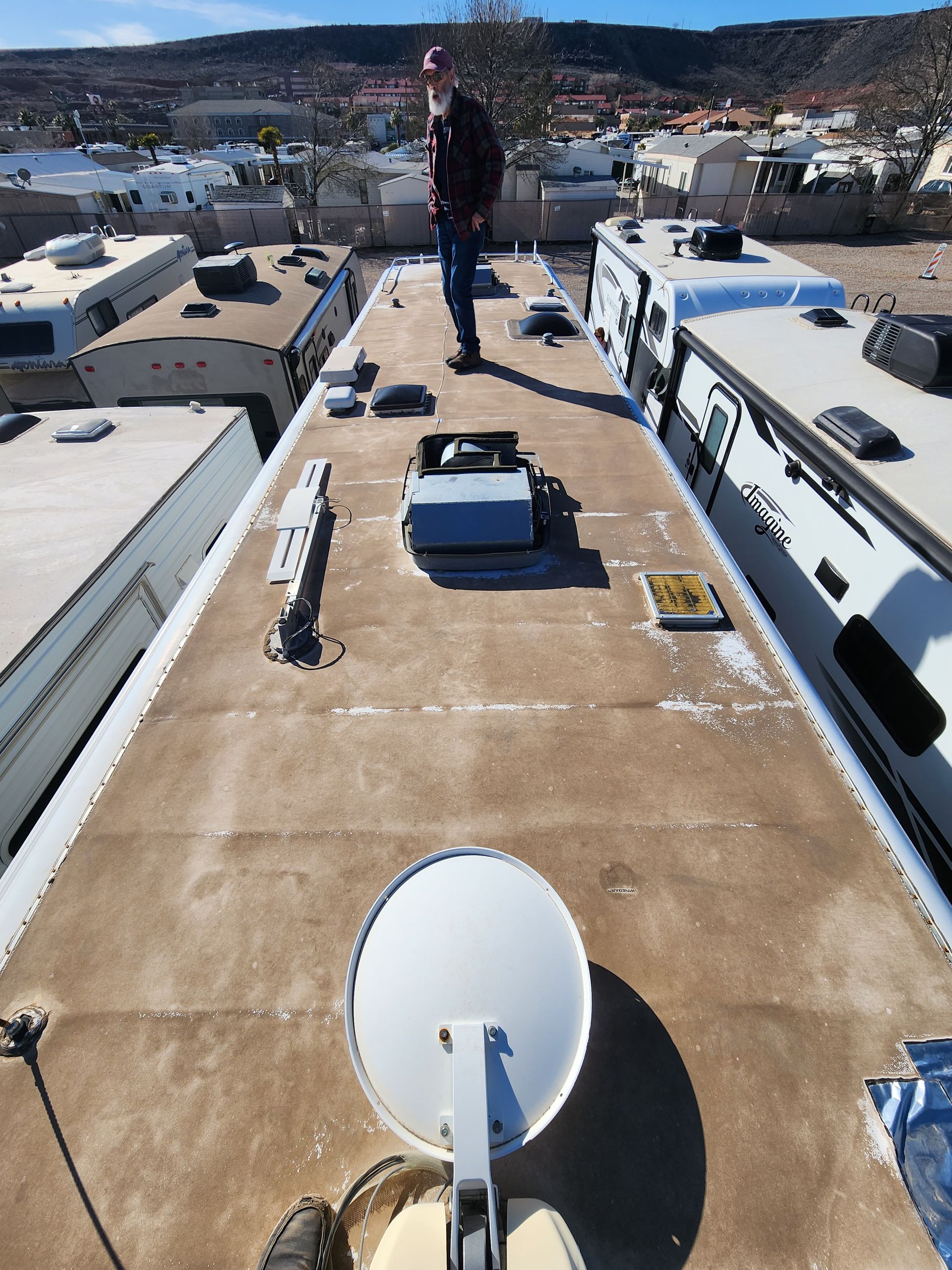 Person stands on RV roof, inspecting equipment. Other RVs are nearby on a sunny day.