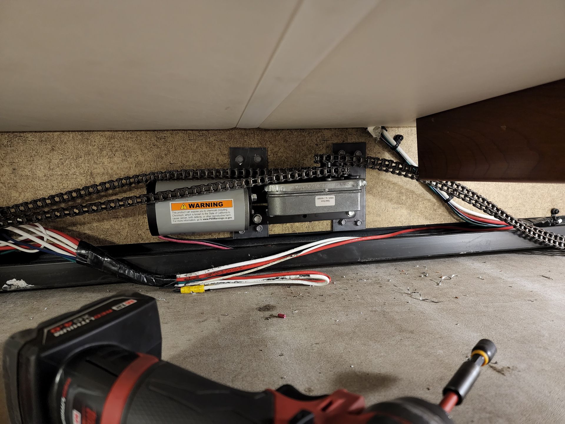 RV slide-out motor and chain assembly. Black metal frame with motors, chains, and wiring. A power tool is at bottom.