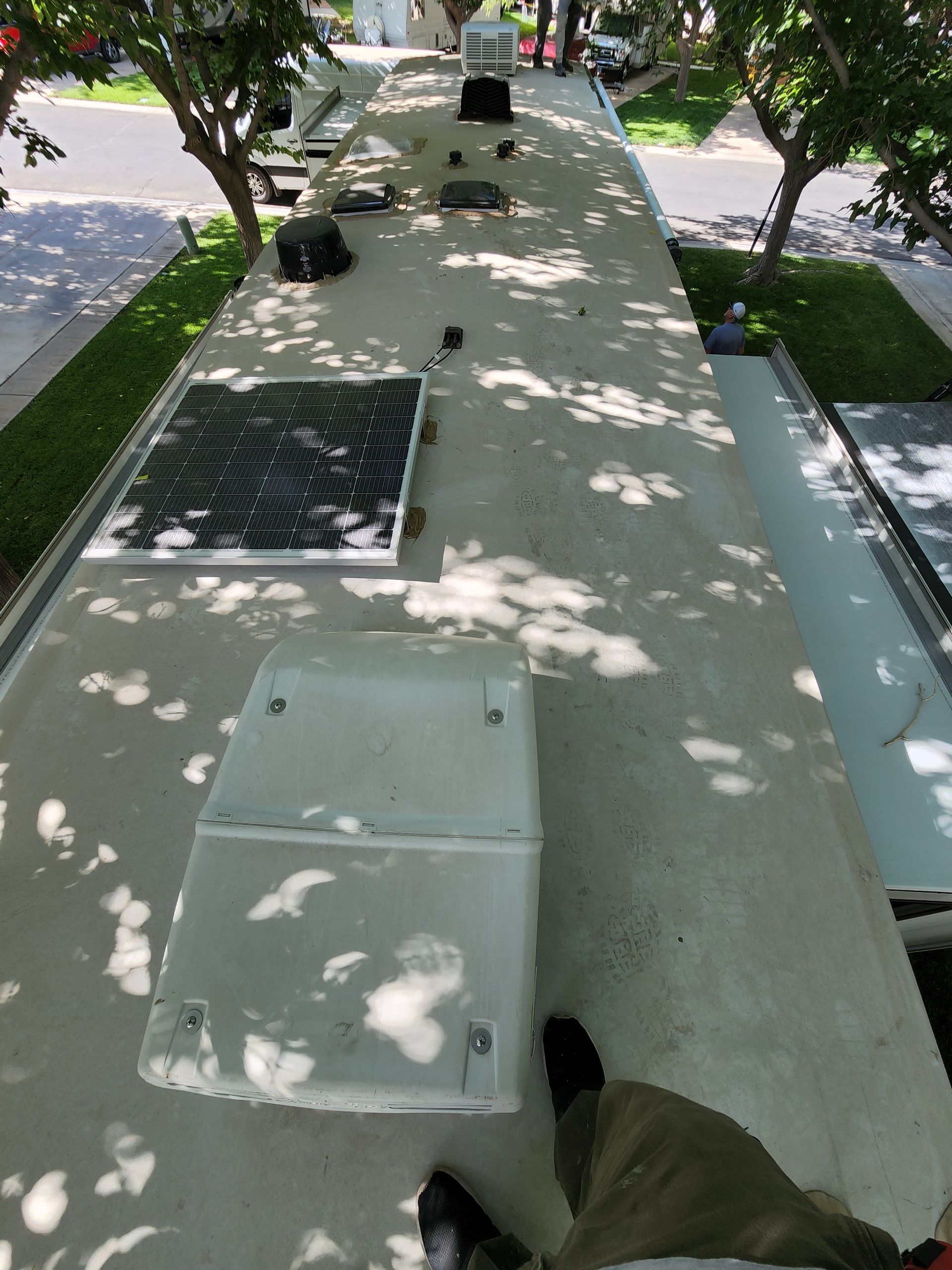 RV roof with solar panels, vents, and air conditioning unit. Gray roof with black panels, sunny outdoors.