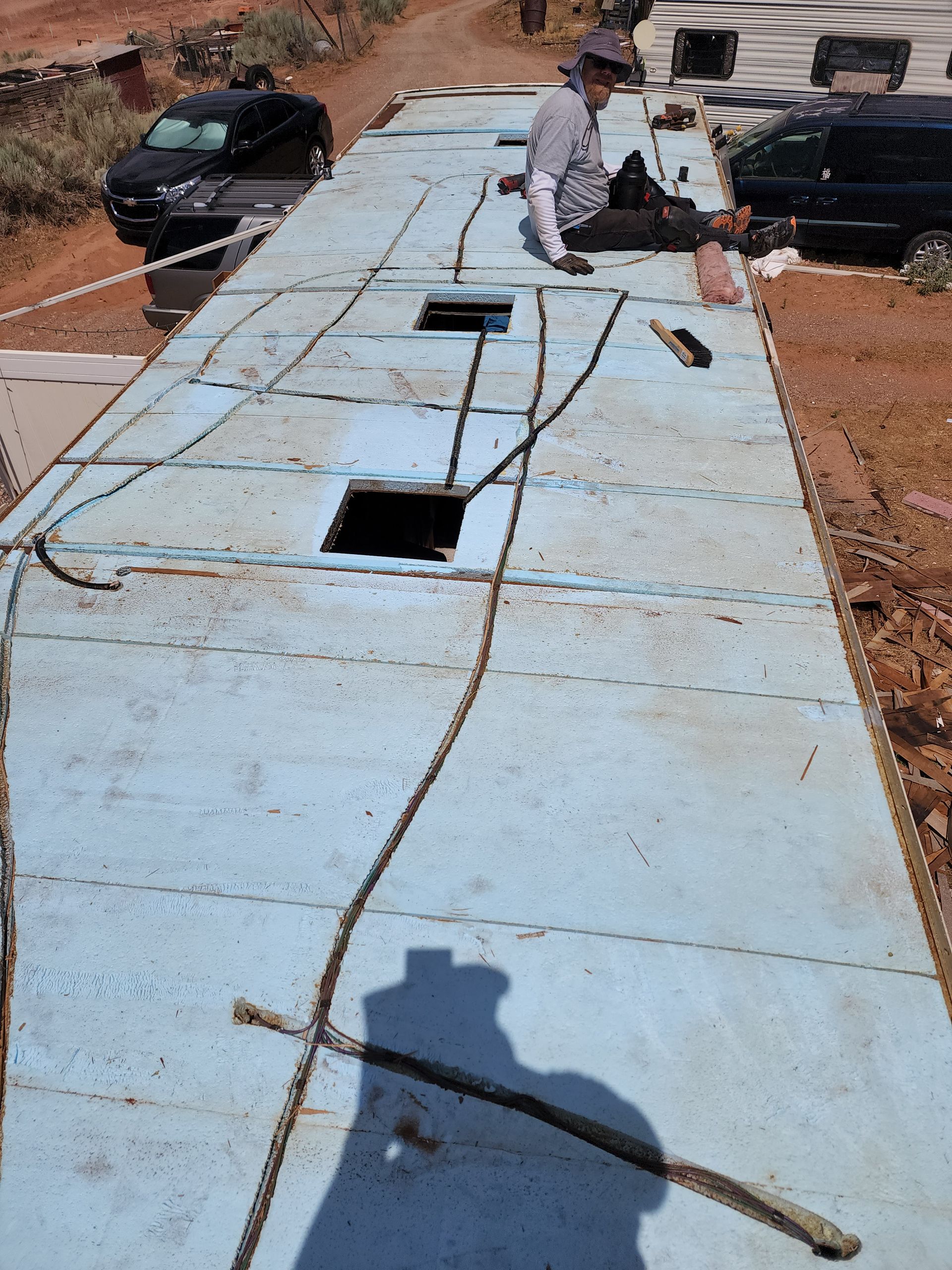 Person working on a long blue-painted structure, likely a roof. Several holes and a car visible.