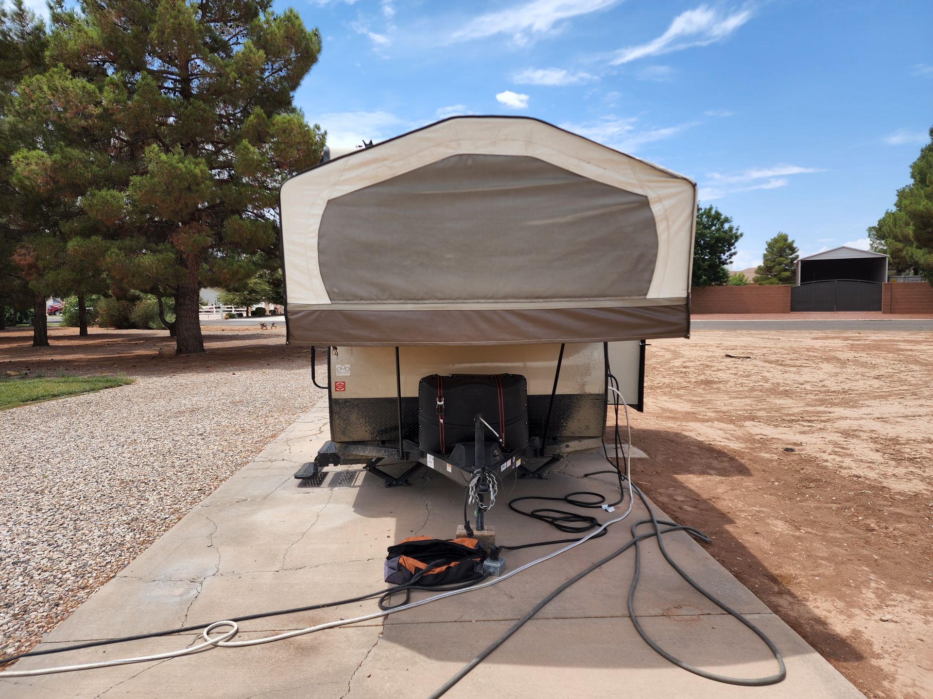Pop-up camper parked on concrete with electrical hookups, set against a backdrop of trees and a blue sky.
