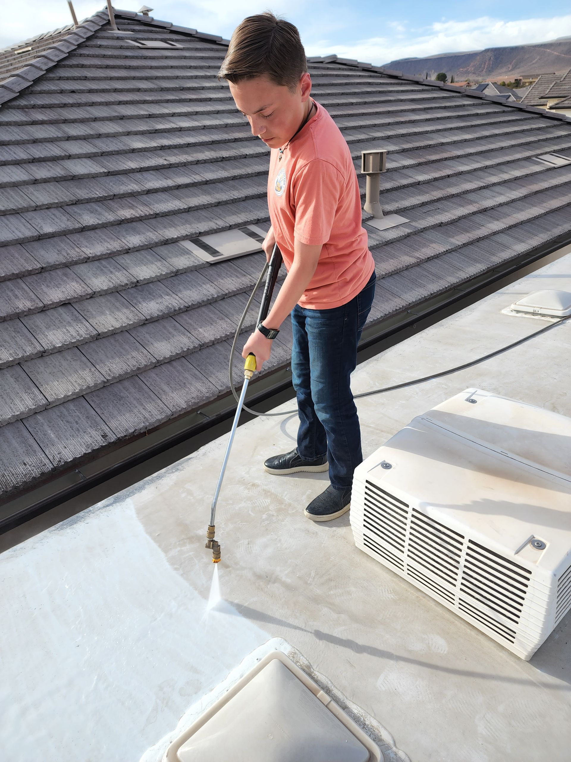Boy sprays a white coating onto a roof with a pressure washer, standing near an air conditioner.