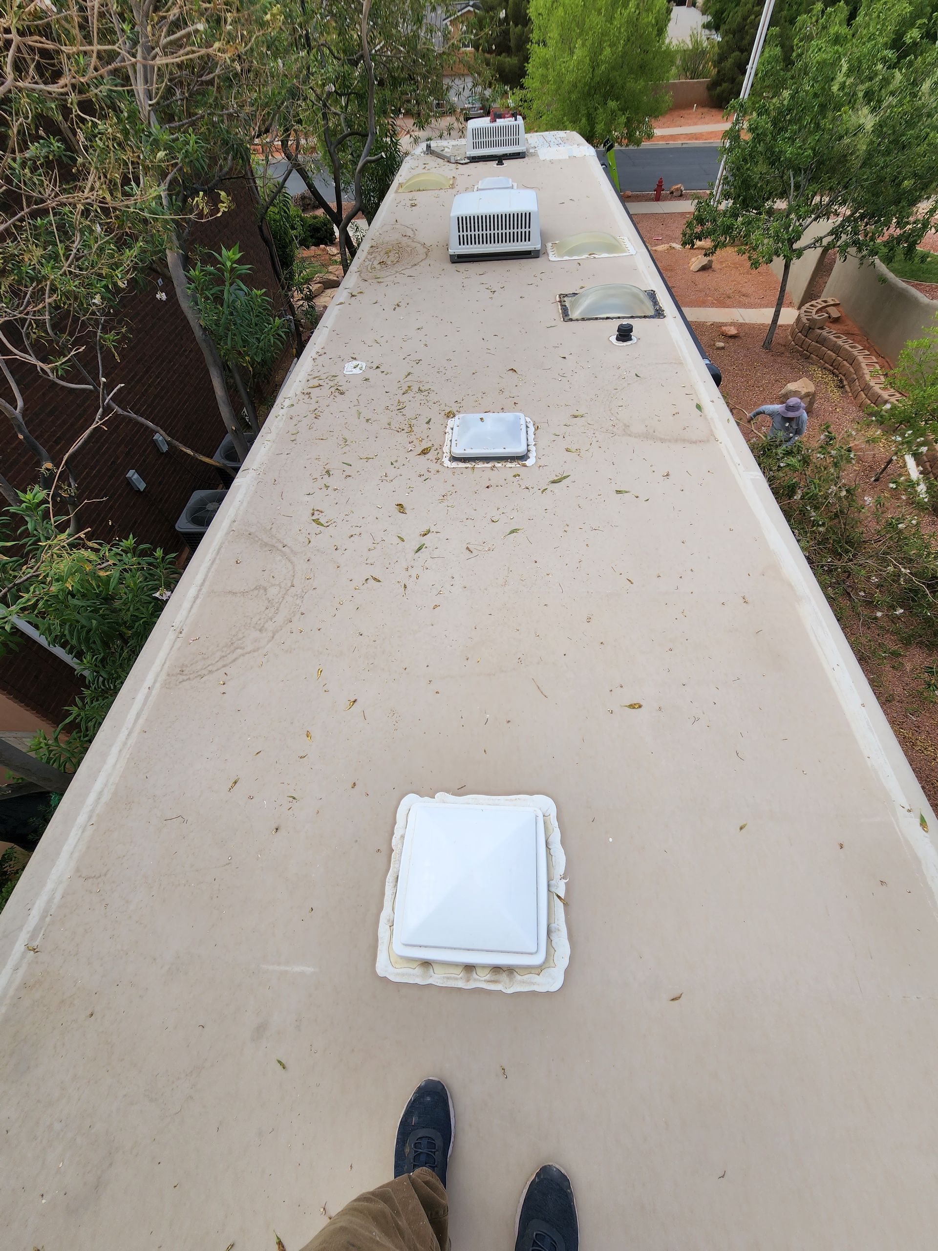 Person standing on RV roof, overview of vents and AC units, trees and buildings in background.