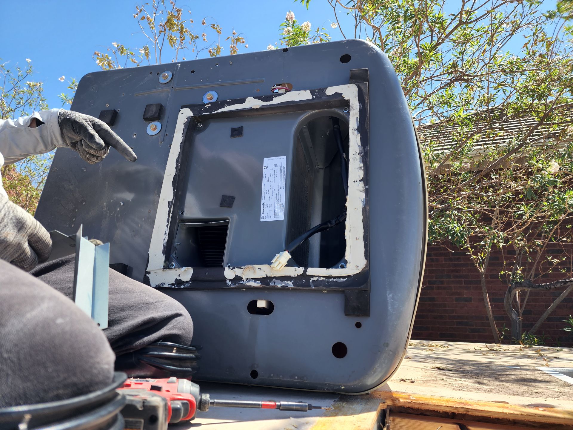 Person working on a gray object outdoors. They are pointing towards a rectangular opening with a white border.