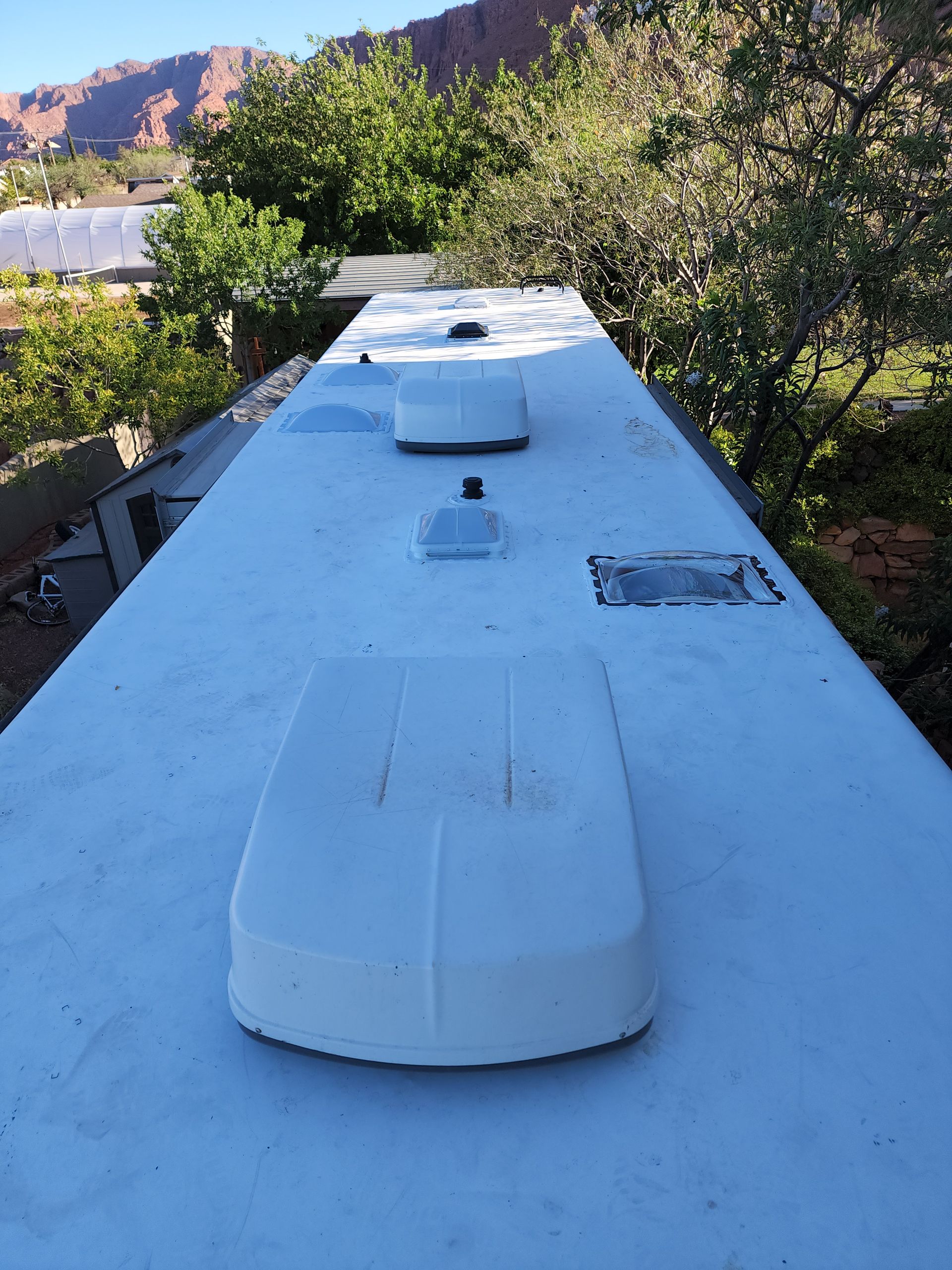 RV roof with air conditioning units and vents, outdoors with foliage and red rock mountains in the background.