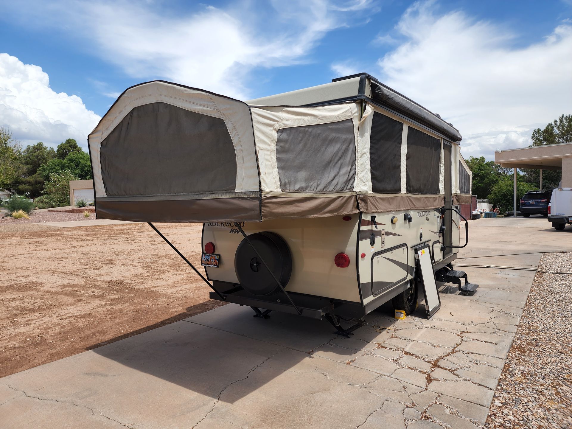 Tan and brown pop-up camper with extended tent sections, parked on a paved driveway with a blue sky.