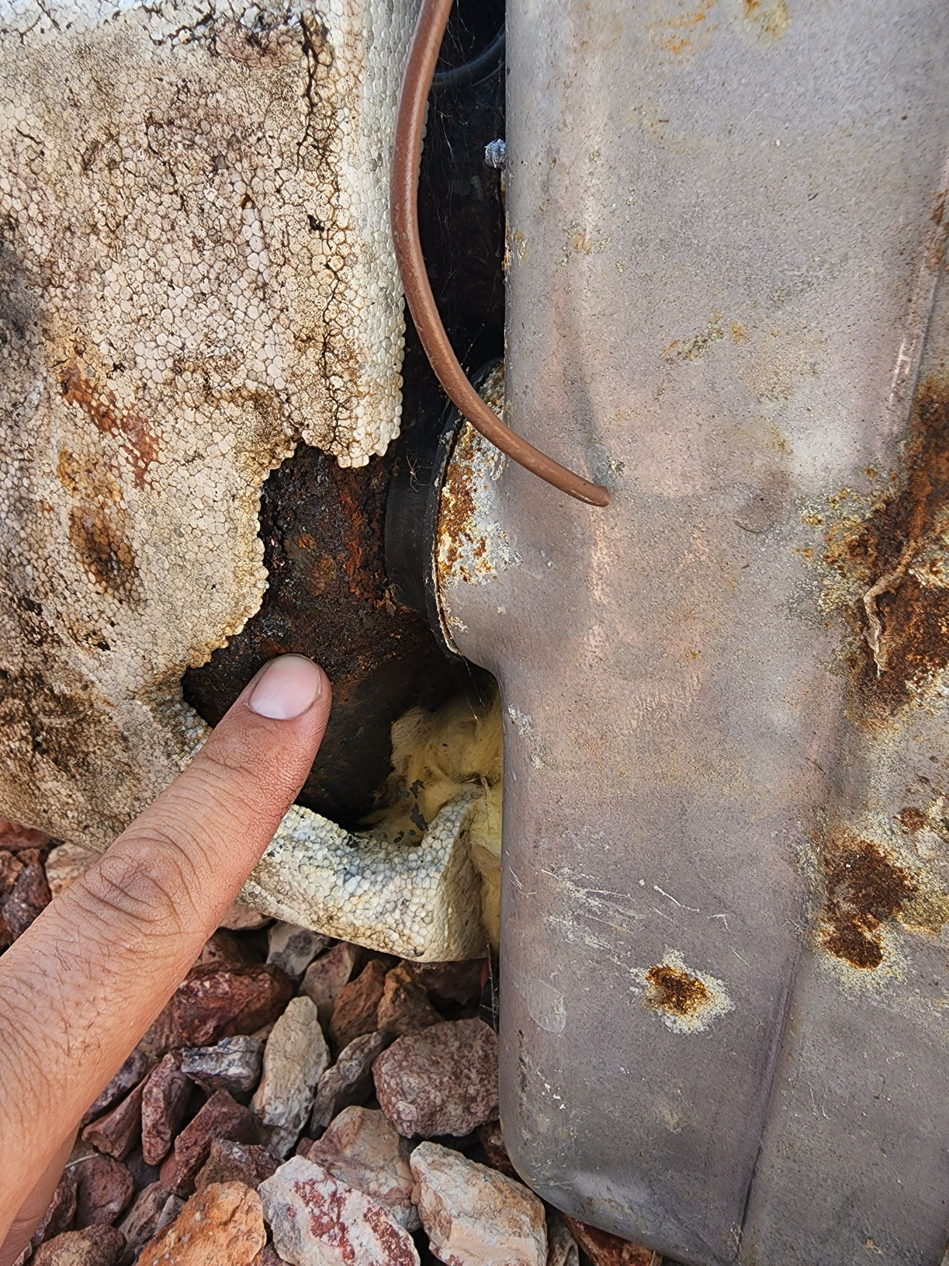 A hand pointing to a corroded metal box with brown rust and a visible hole filled with yellow insulation.