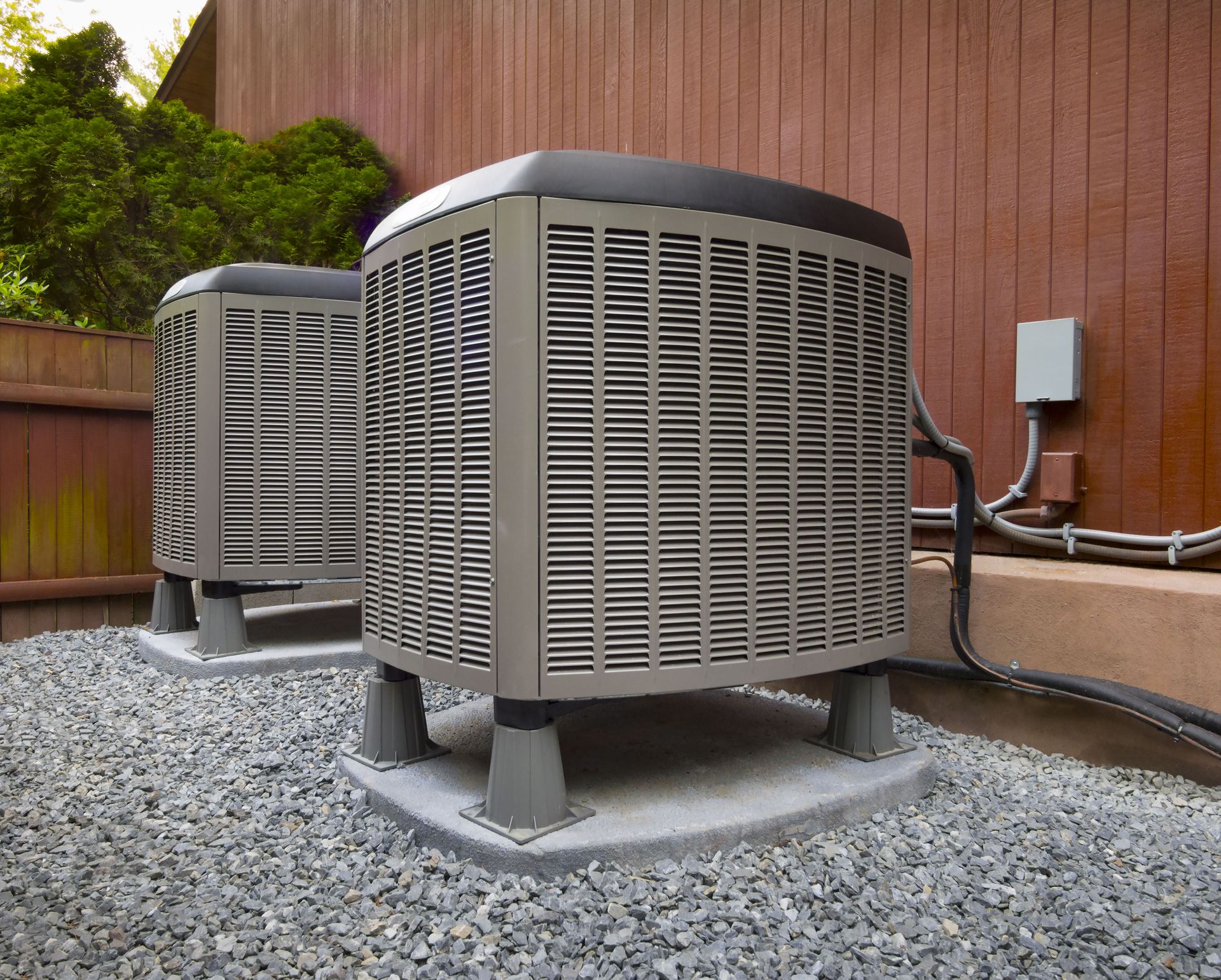 Two air conditioning units on gravel next to a brown wooden fence.