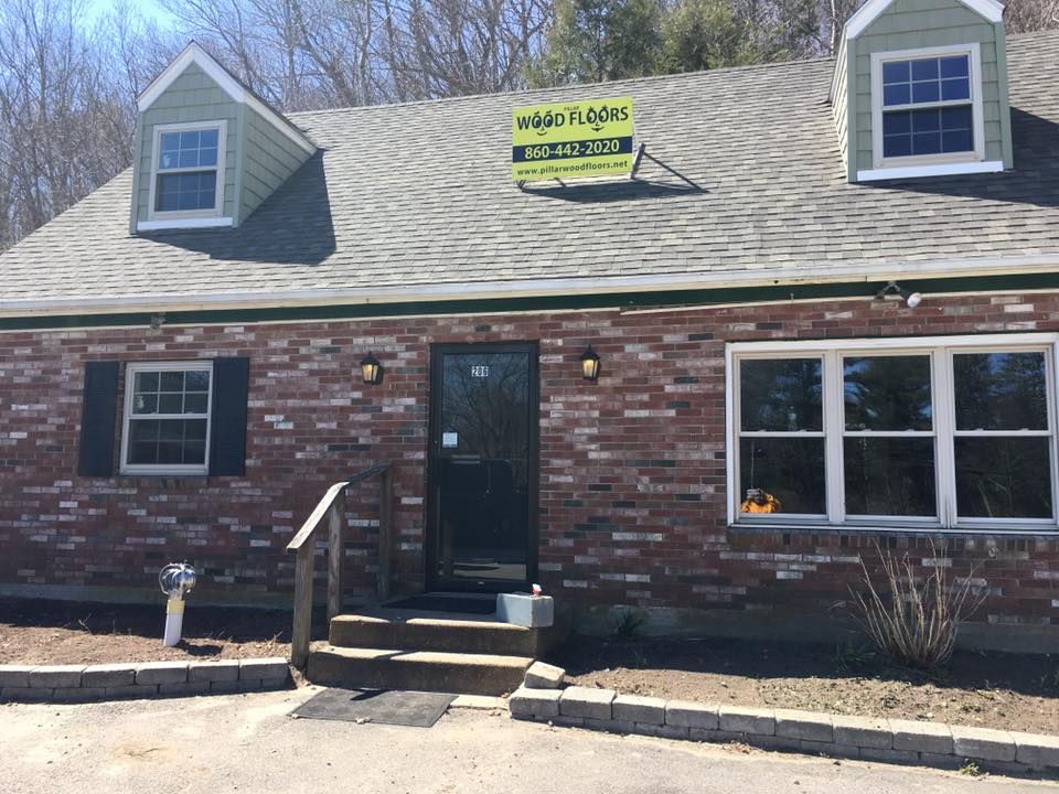 Brick building with a black door, windows, and a sign on the roof; flowers and a small walkway in front.