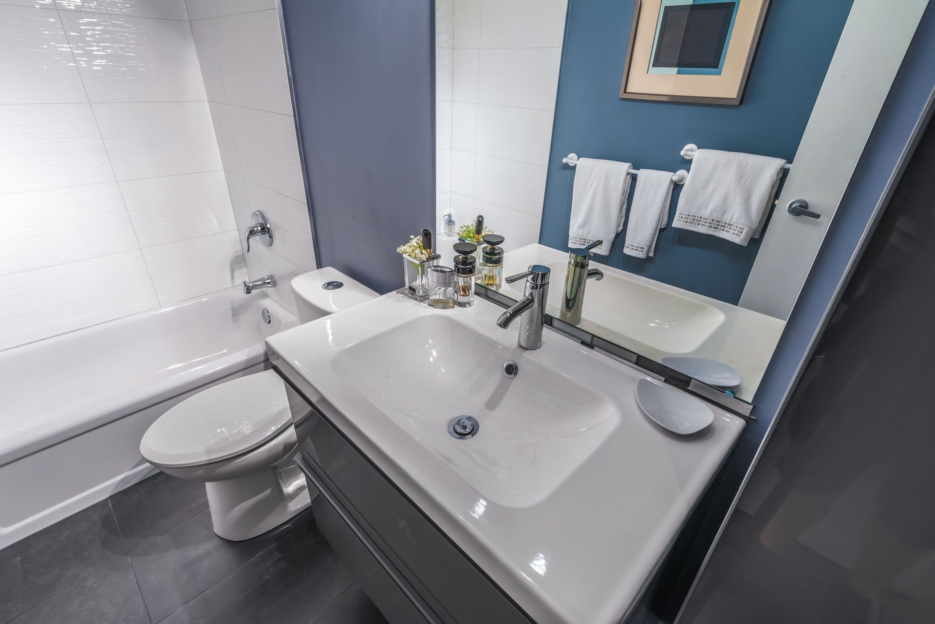 Modern bathroom with white sink, toilet, and bathtub. Blue and white walls, dark floor, towels.