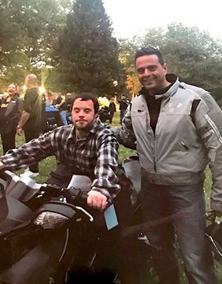 Man on motorcycle with friend; smiling outdoors. One in plaid shirt, other in motorcycle gear.