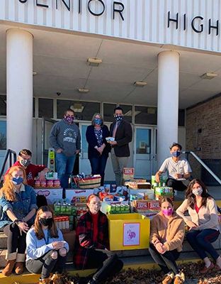 People in front of a school, posing with donations. Canned goods, boxes, and a check are visible.