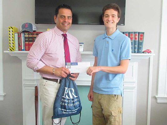 Man in pink shirt hands a check and bag to a teen in blue shirt, in front of a fireplace.