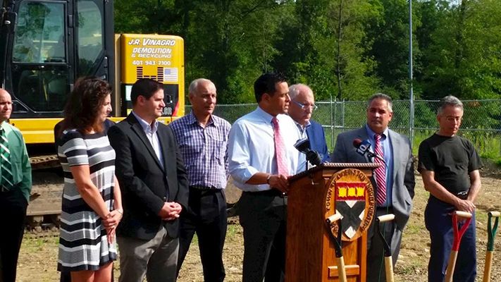 People at a podium with a logo, ground-breaking ceremony. Backhoe in background.