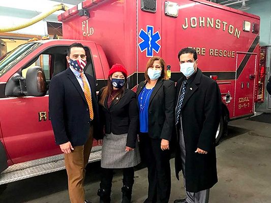 Four people pose in front of a red ambulance. They wear masks and formal attire. The ambulance reads 