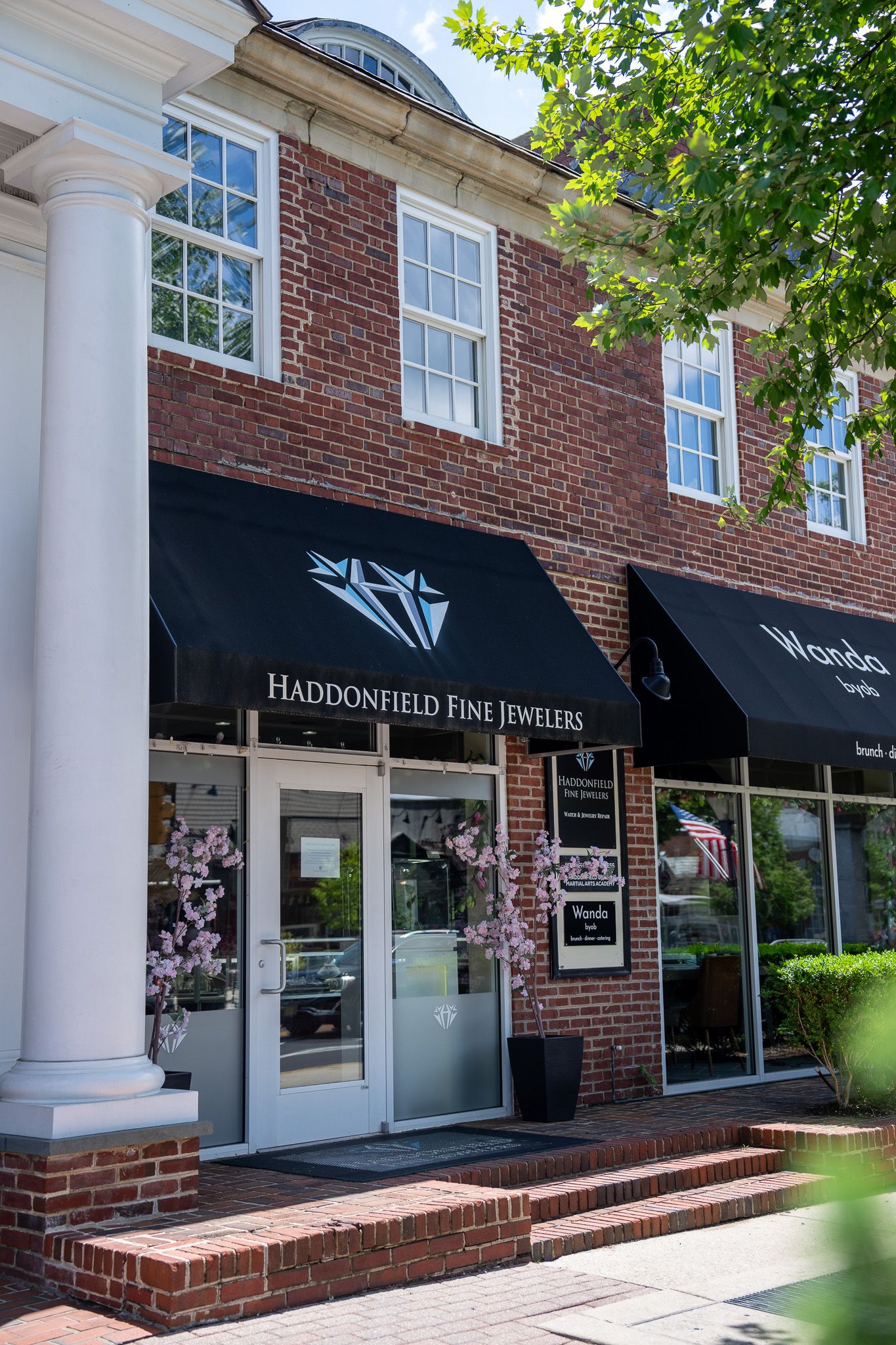 Brick storefront with black awnings and a glass door, framed by white columns and greenery