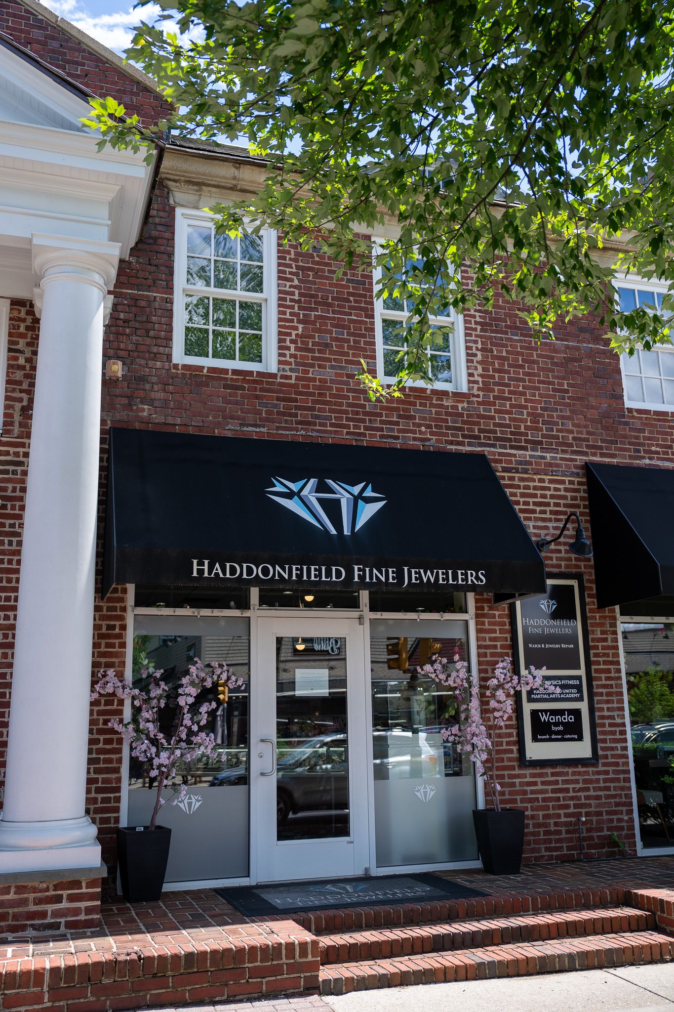 Brick storefront with black awning and glass doors, framed by white columns and leafy trees.