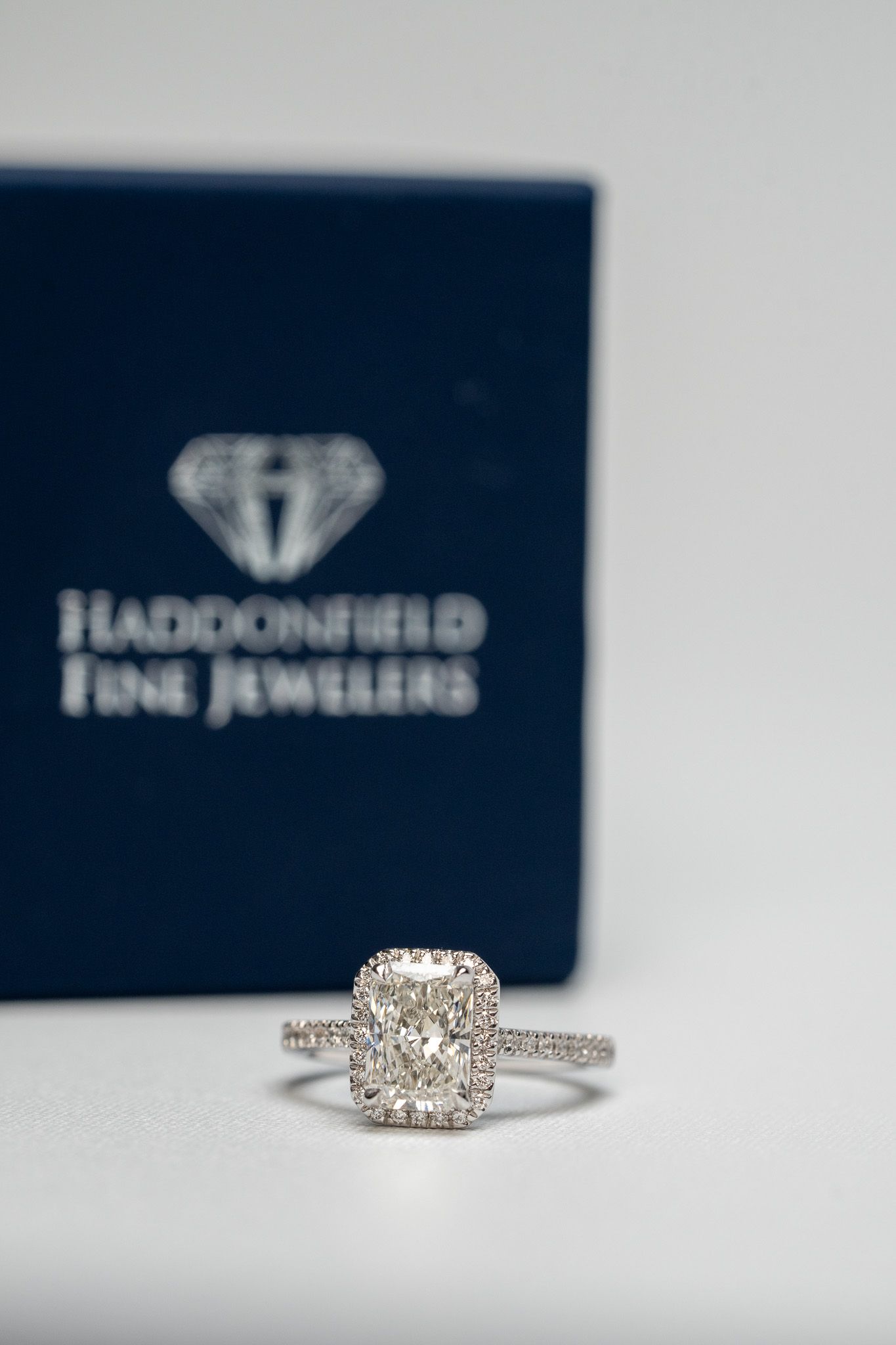 Diamond engagement ring in front of a blurred black jewelry box with a logo and text.