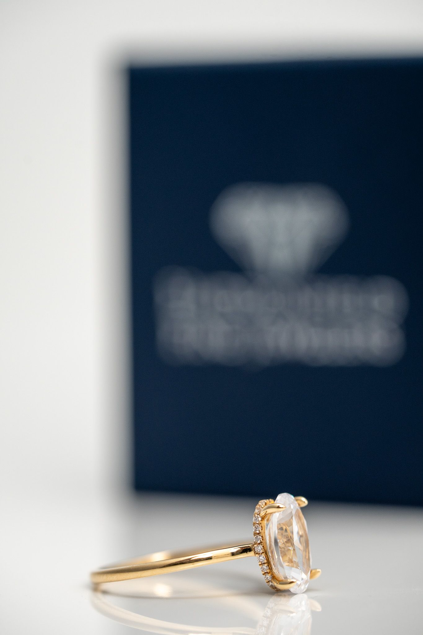Gold ring with diamond on white surface, with blurred navy jewelry box in the background