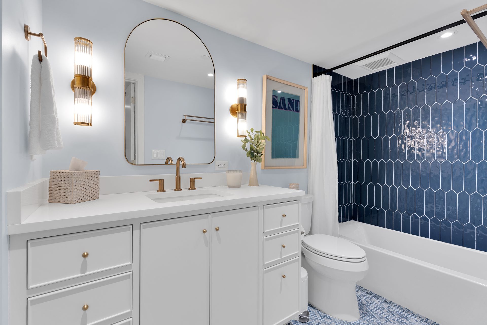 Bright white bathroom with gold fixtures, arched mirror, blue shower curtain, and patterned floor tile