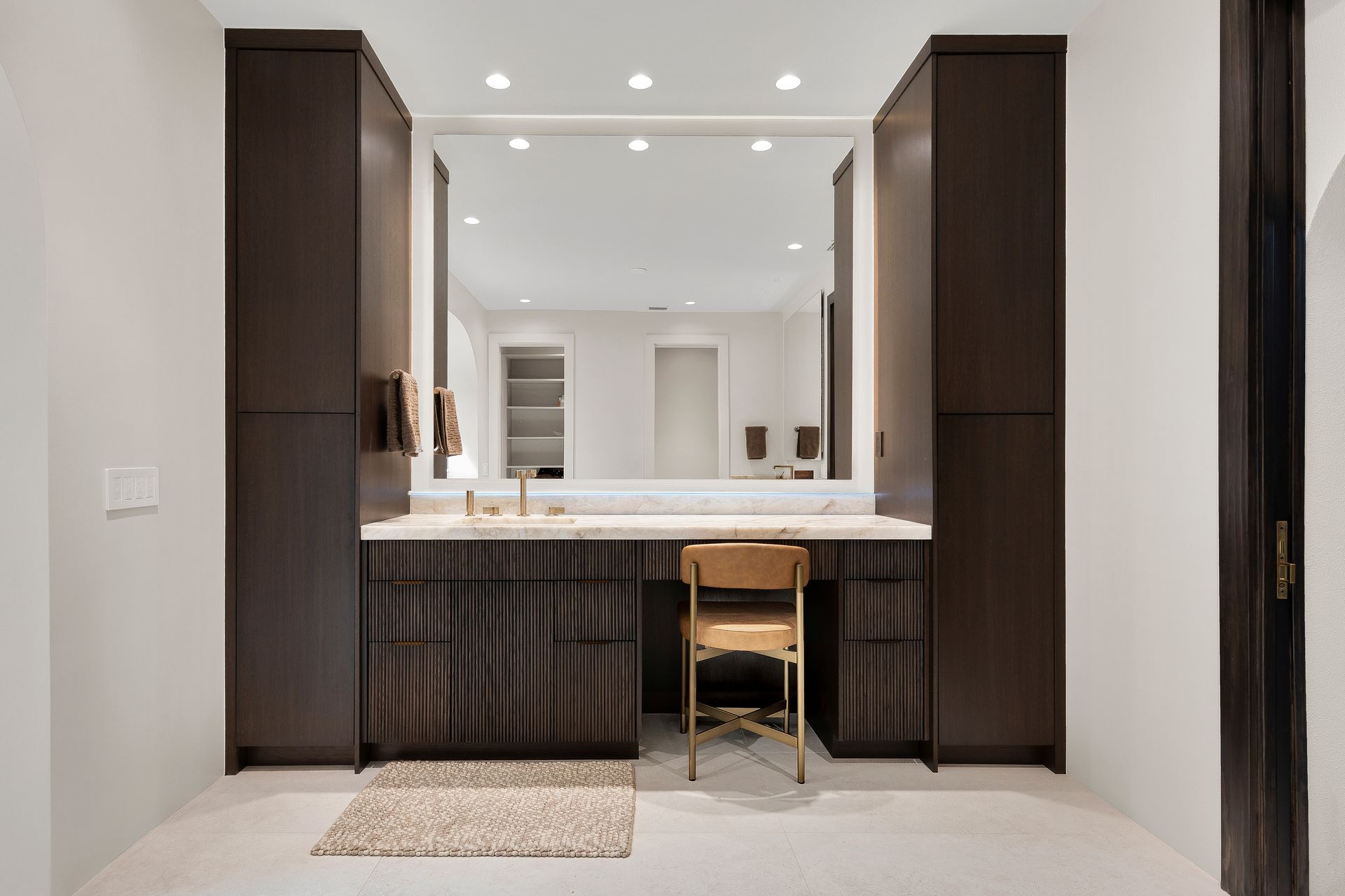 Modern bathroom vanity with dark wood cabinets, large mirror, white countertop, and a stool centered below.