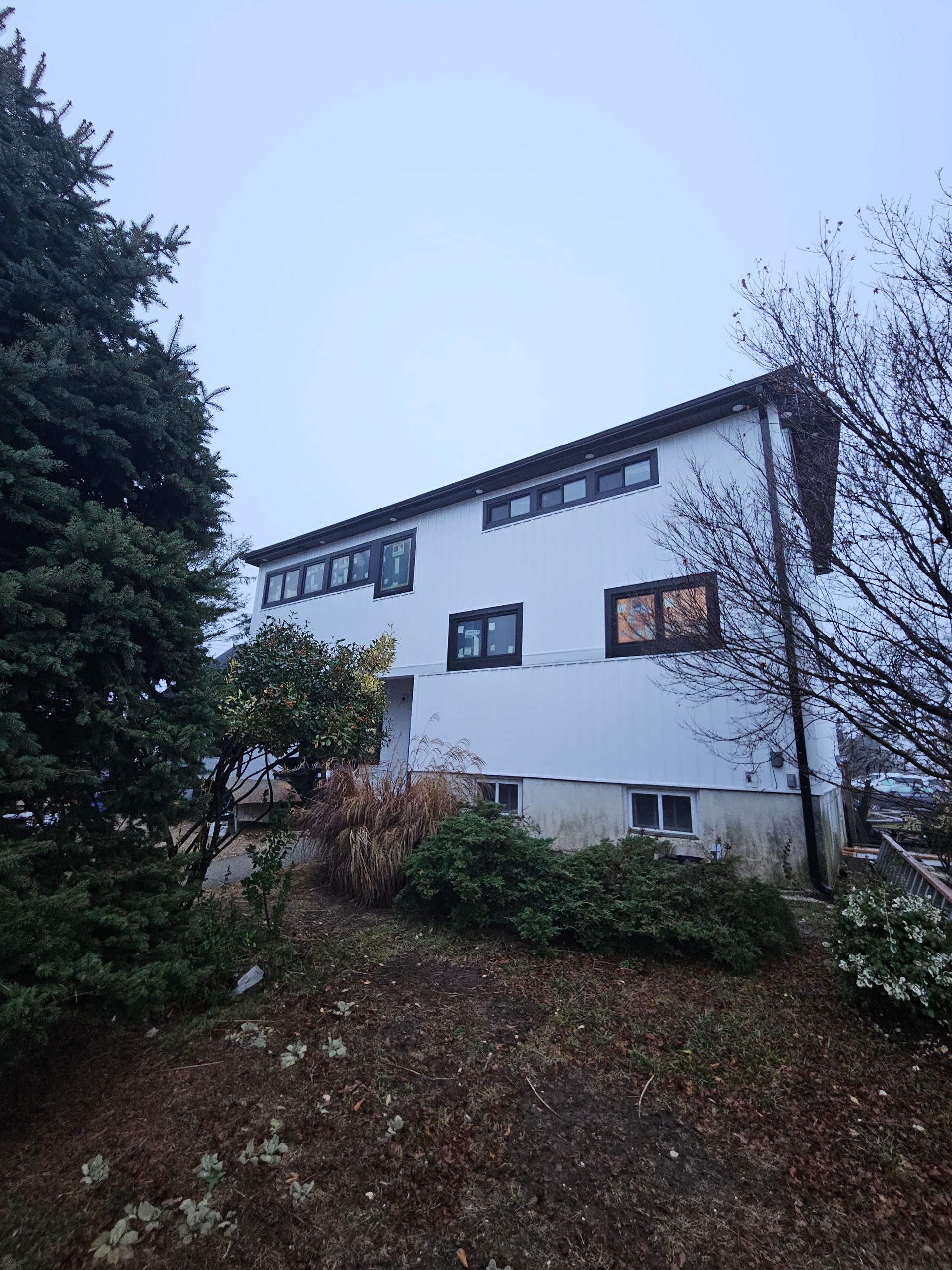 White two-story house with dark-framed windows, set on a hillside with trees and shrubs against a cloudy sky.